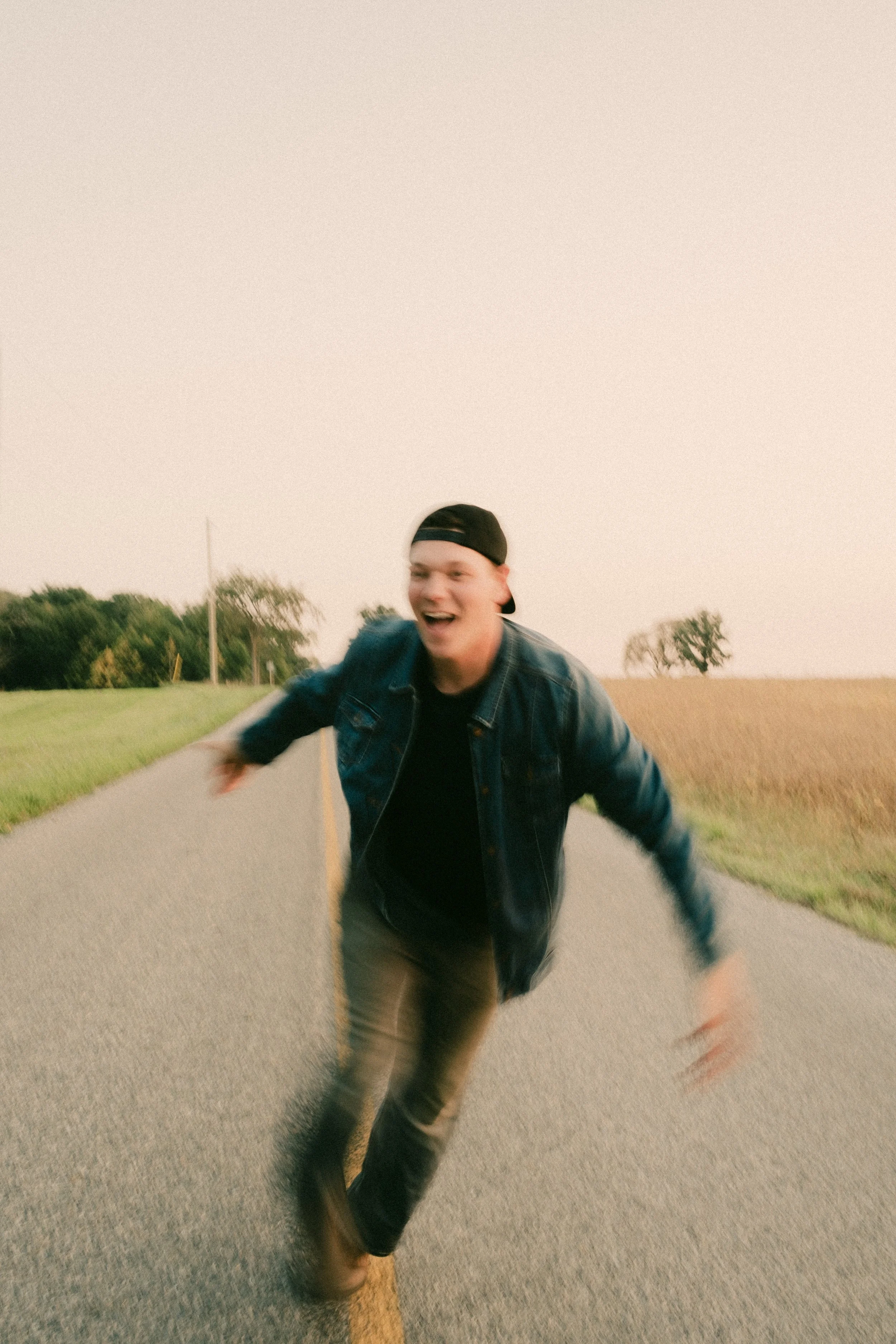 A young man runs toward the camera on an empty rural road with open fields on either side, smiling and laughing with arms outstretched.