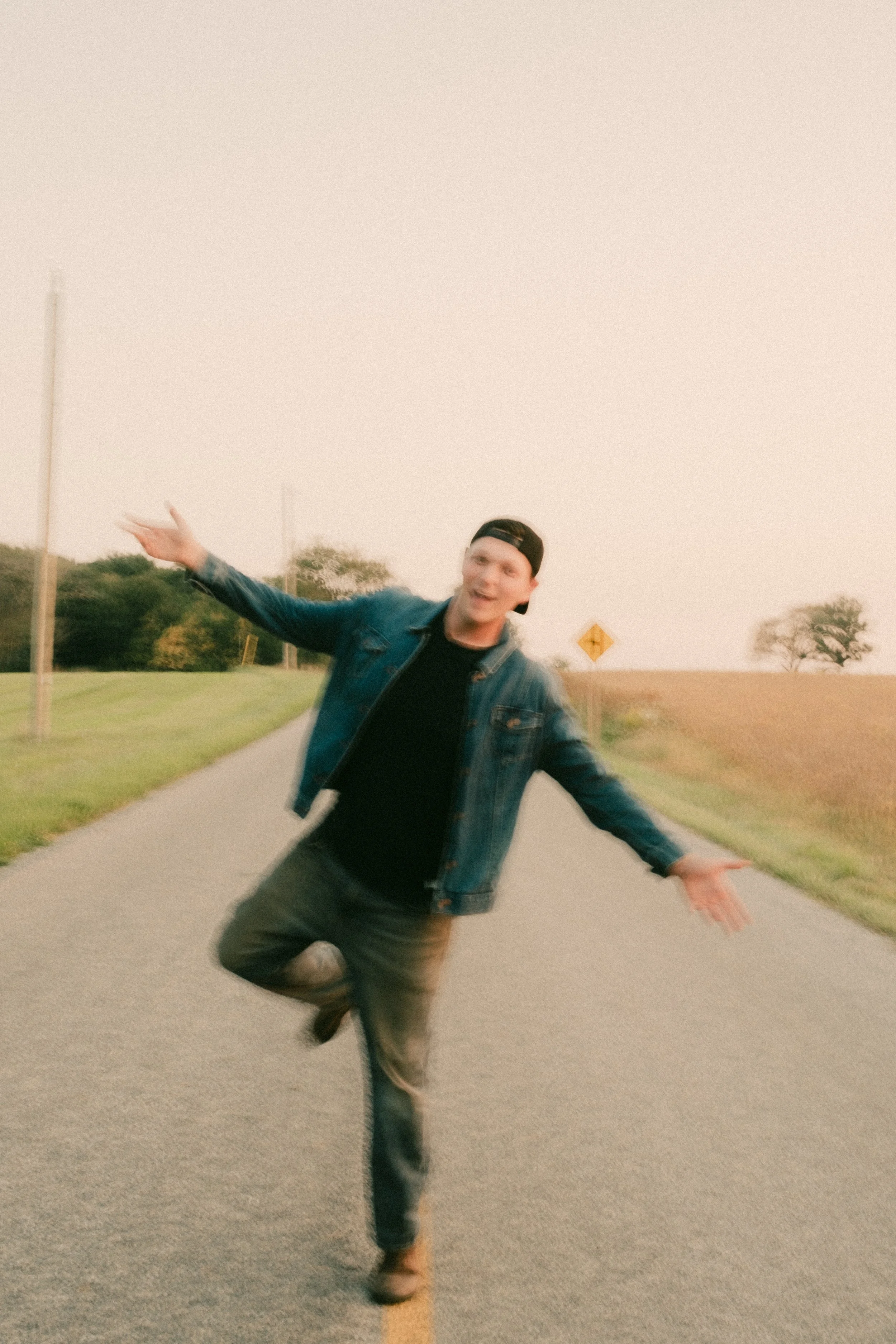 A young man in a denim jacket and black cap standing on a rural road with arms outstretched and one leg lifted, smiling.