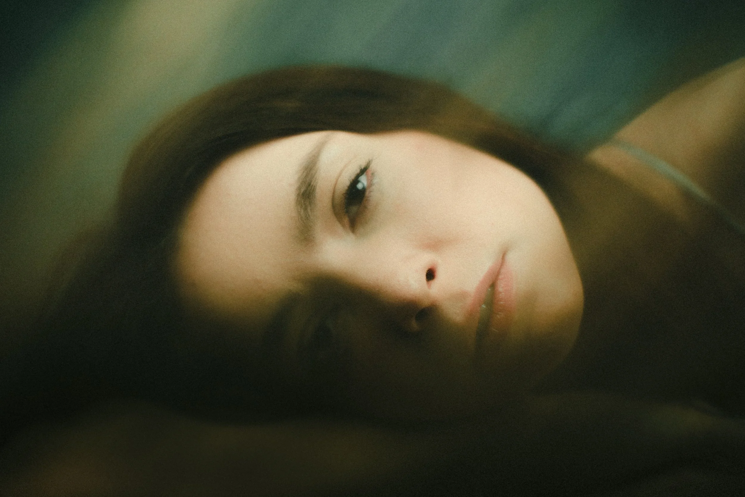 A young woman with long brown hair lying down, with her head resting on a dark surface. She has a calm expression, light skin, and subtle makeup.