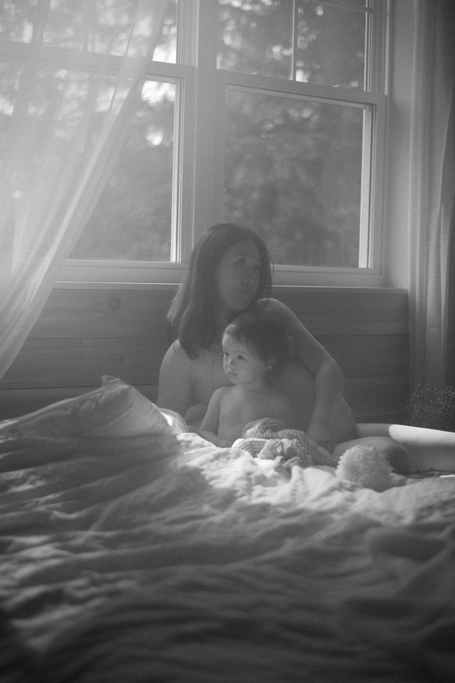 A woman with shoulder-length hair and a young child sitting on a bed near a window, both looking out, with sunlight coming through the curtains. Black and white photo.