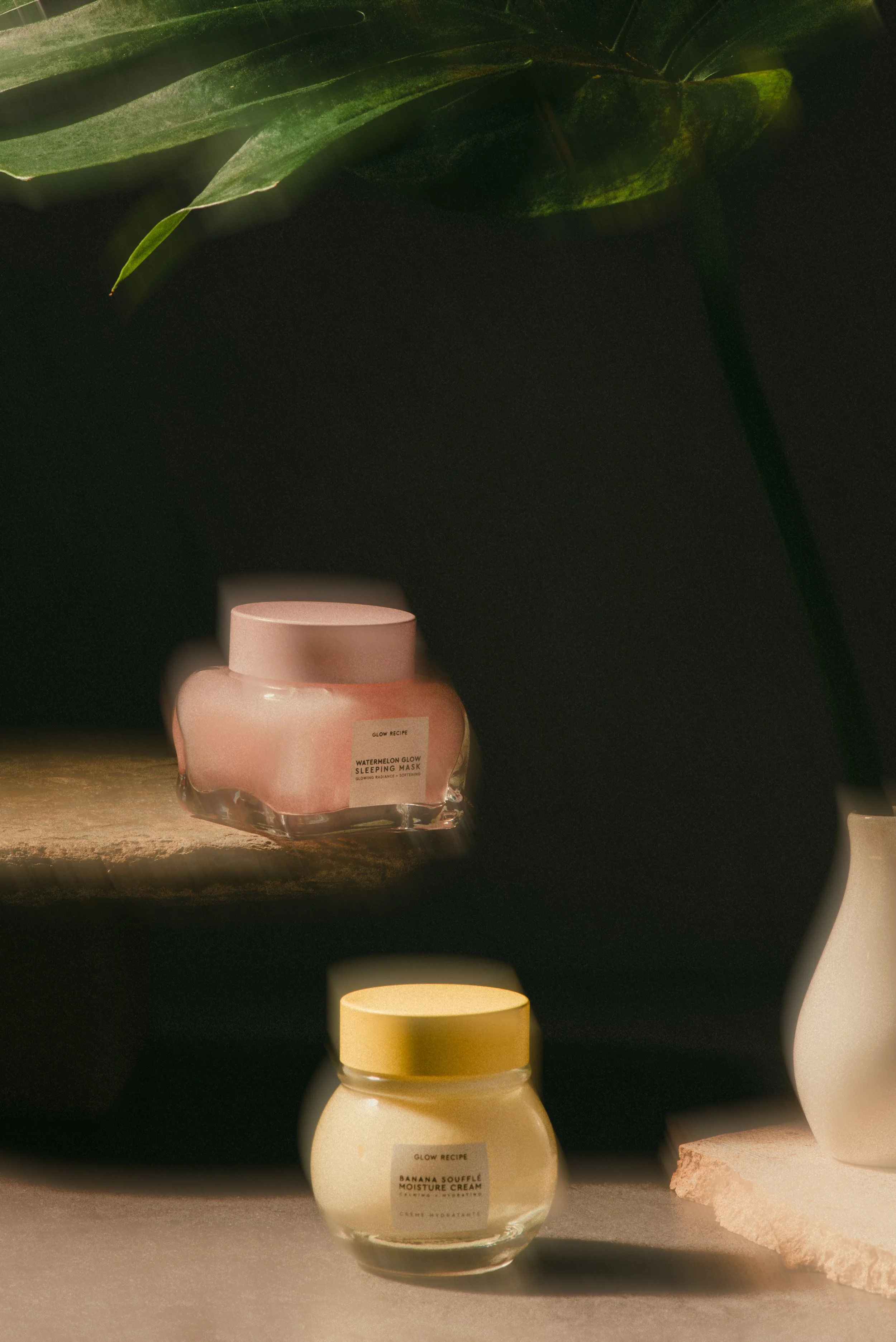 Two jars of skincare cream, one pink labeled 'Watermelon Glow Sleeping Mask' on a rock shelf, the other yellow labeled 'Banana Soufflé Moisture Cream' on a concrete surface, with green leaves and a dark background.