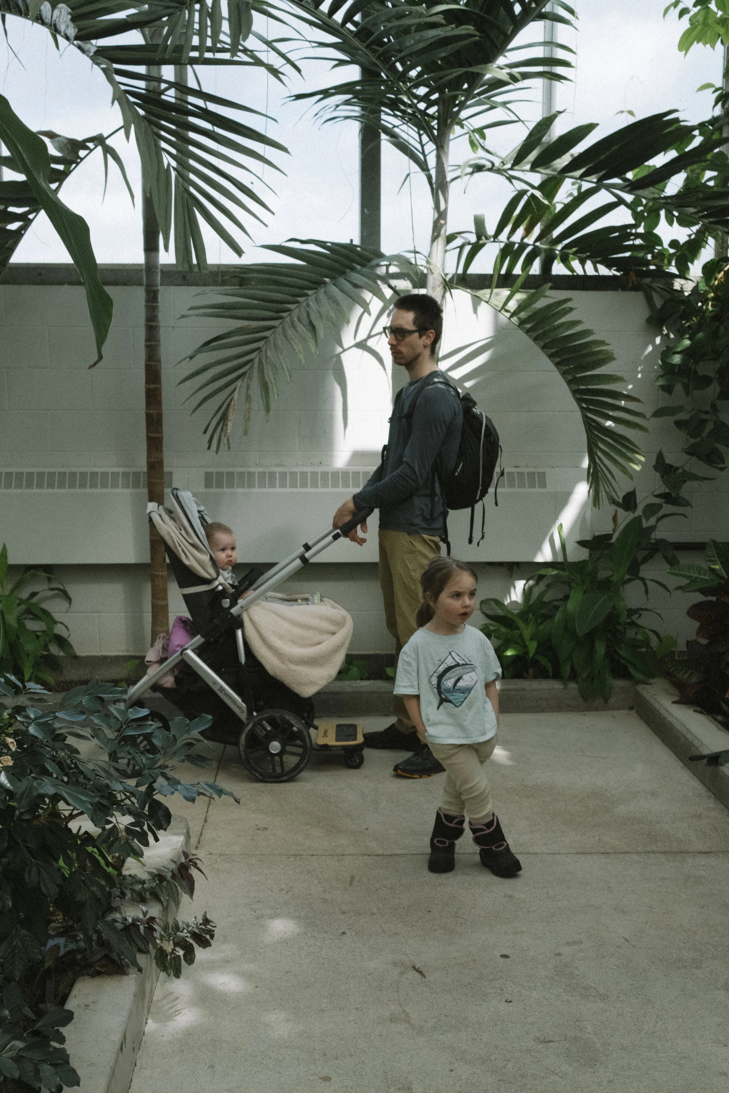 A man with a backpack walking through an indoor botanical garden, pushing a stroller with a young child inside, while a girl with a gray t-shirt and beige pants stands nearby.