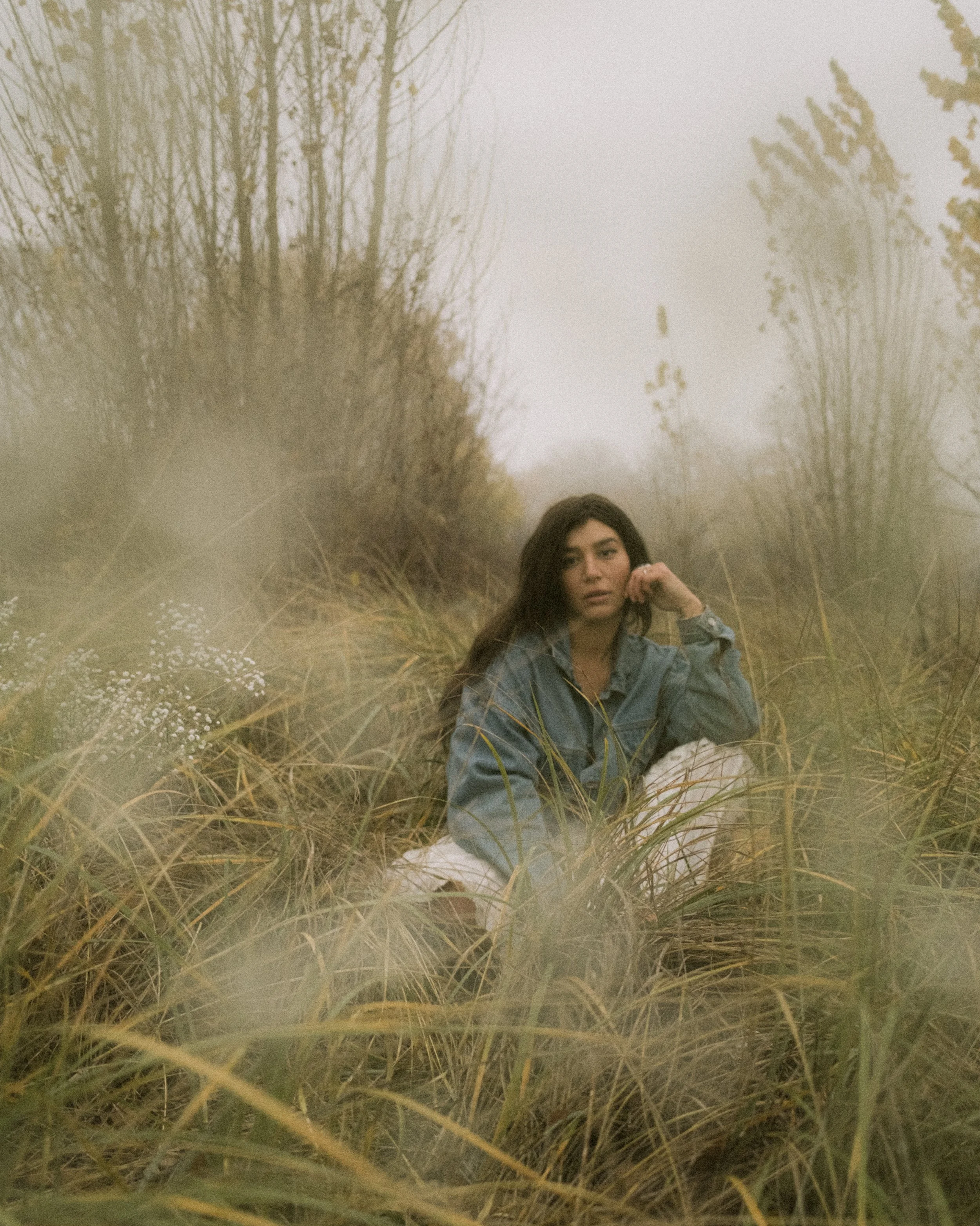 Outdoor portrait of a young woman in denim and white skirt, sitting in tall grass with fog and bare trees, evoking a moody, ethereal atmosphere