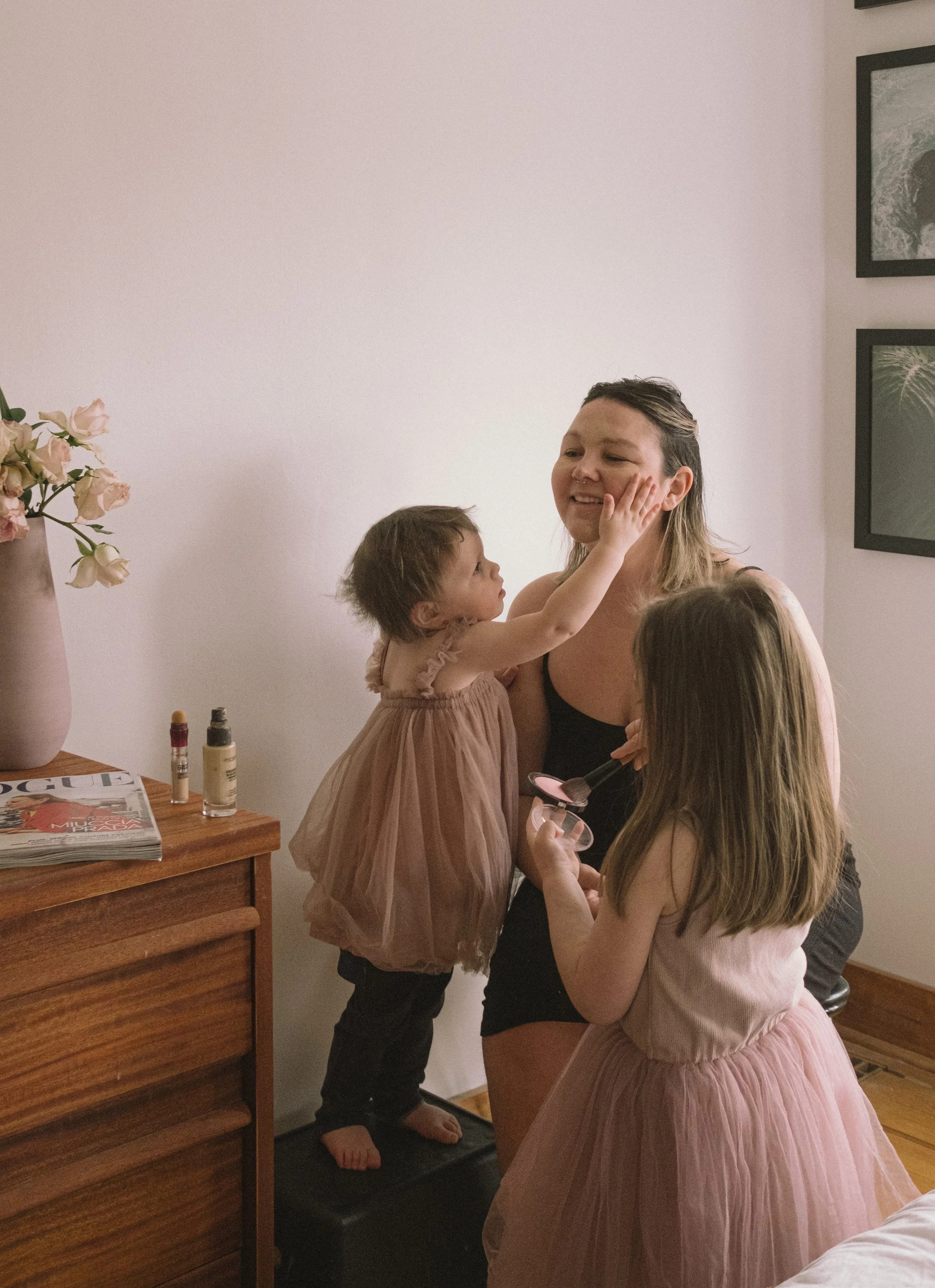 A woman having makeup applied by two young girls in a bedroom with artwork on the wall and a table with magazines and bottles on it.