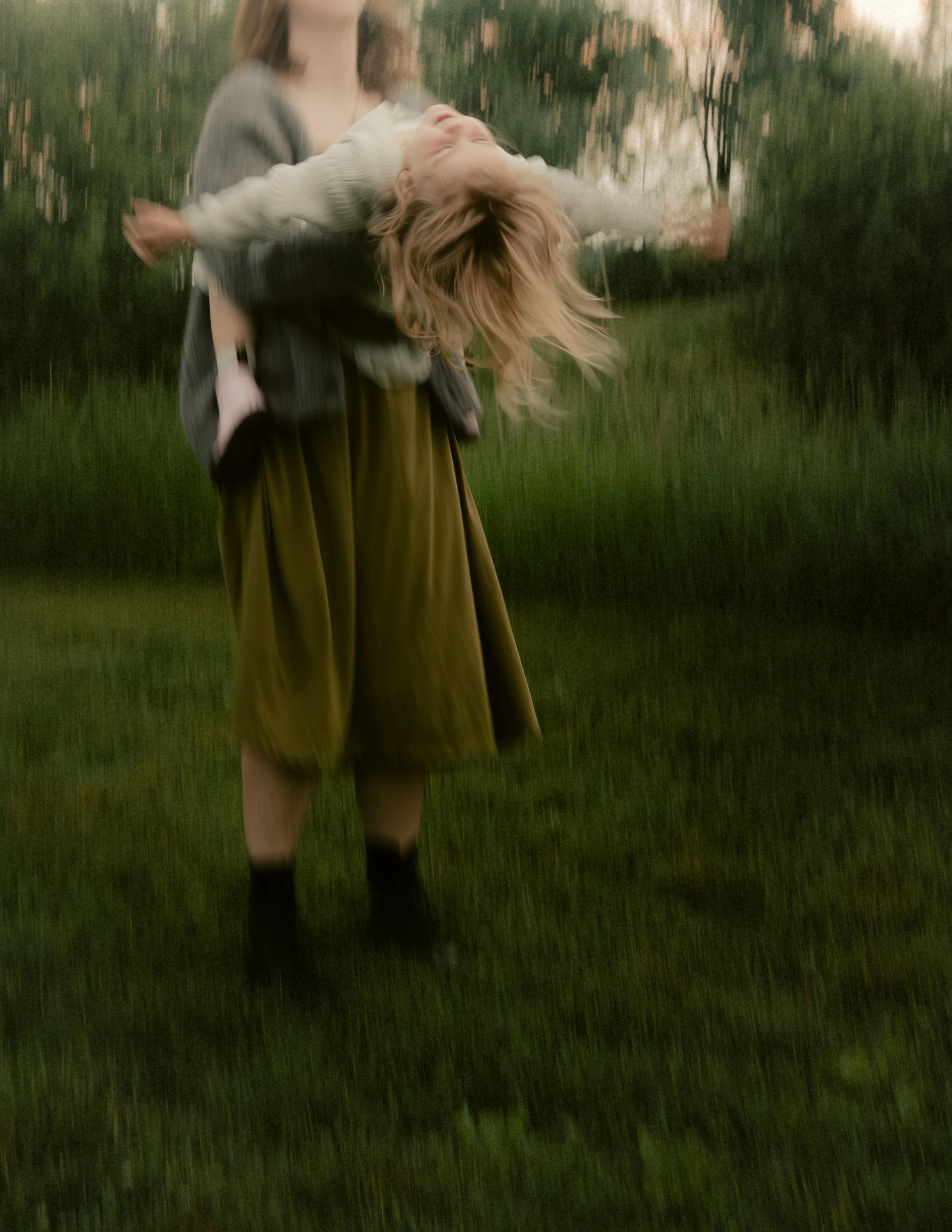 A woman holding a girl upside down in a grassy outdoor area during sunset, with trees in the background.