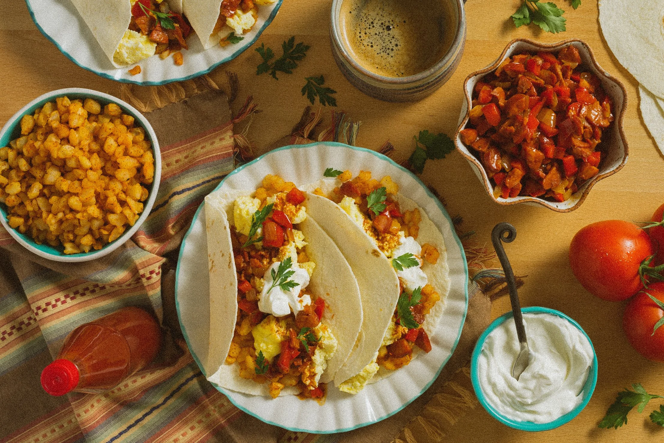 Tacos with filling, bowls of corn and vegetable salsa, tomatoes, sour cream, and a glass of beer on a wooden table.