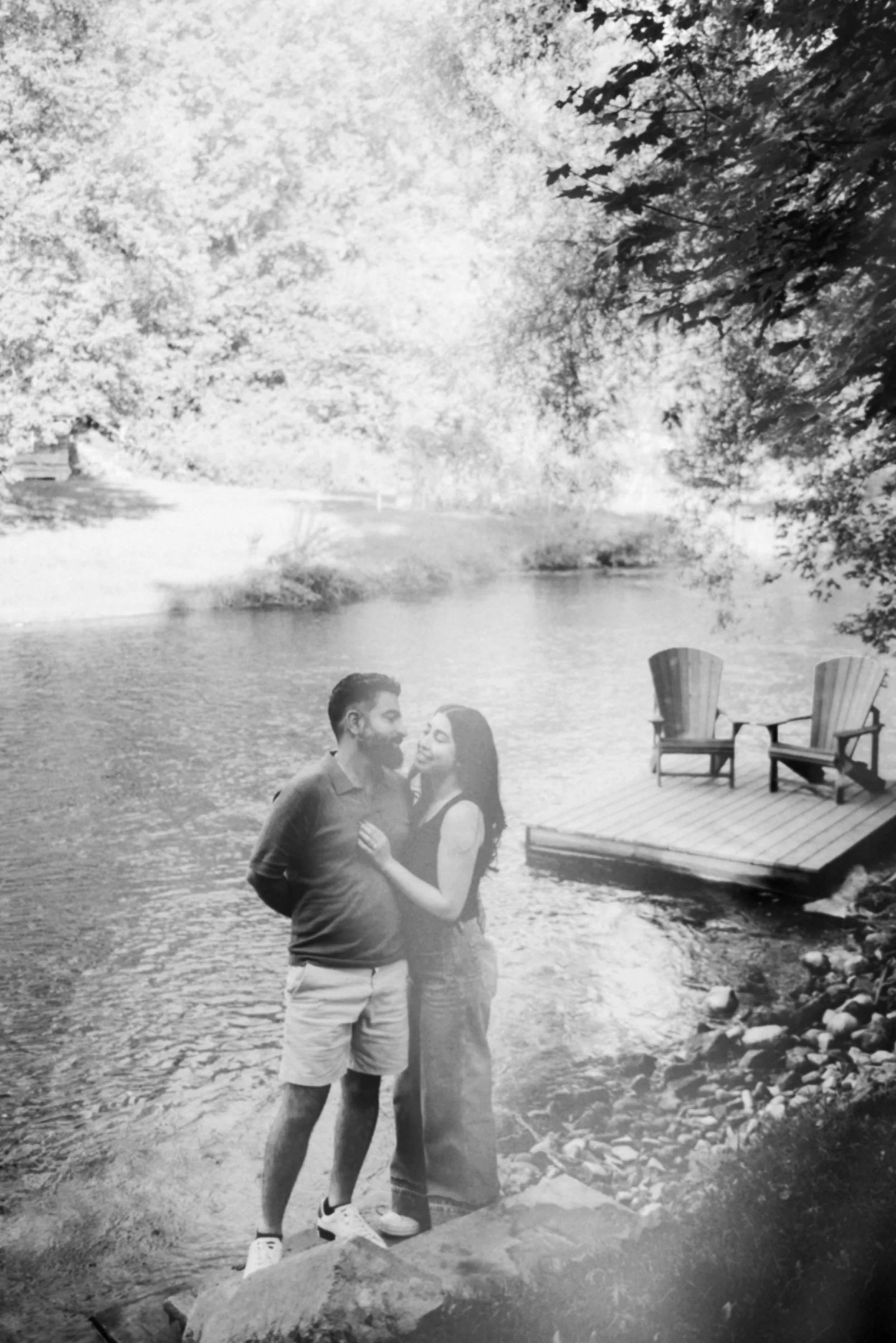 A black and white photograph of a couple standing by a river, with trees and a wooden deck with two chairs in the background.