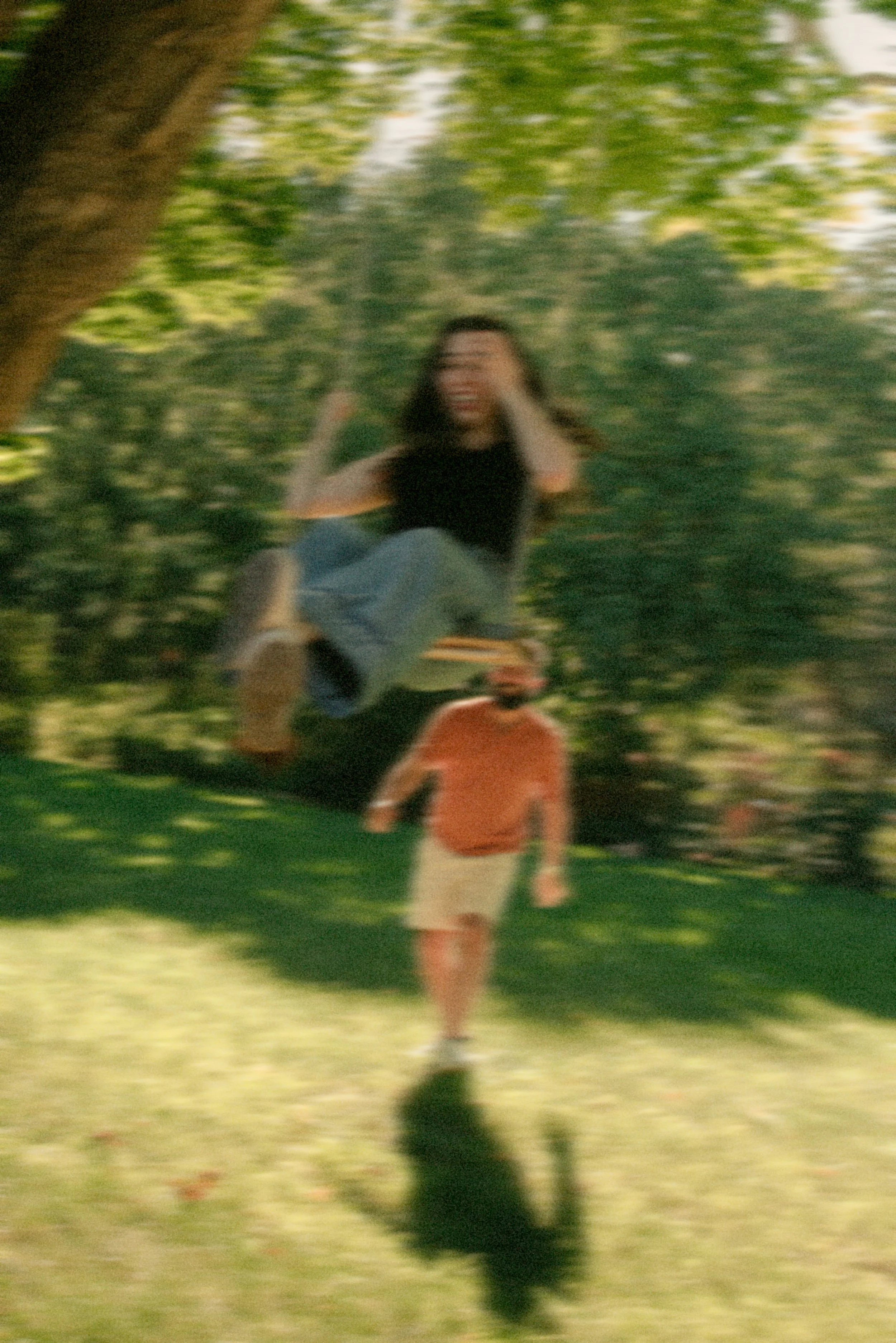 A woman on a swing in a park, with a man pushing her on grass in the background, trees and greenery around.