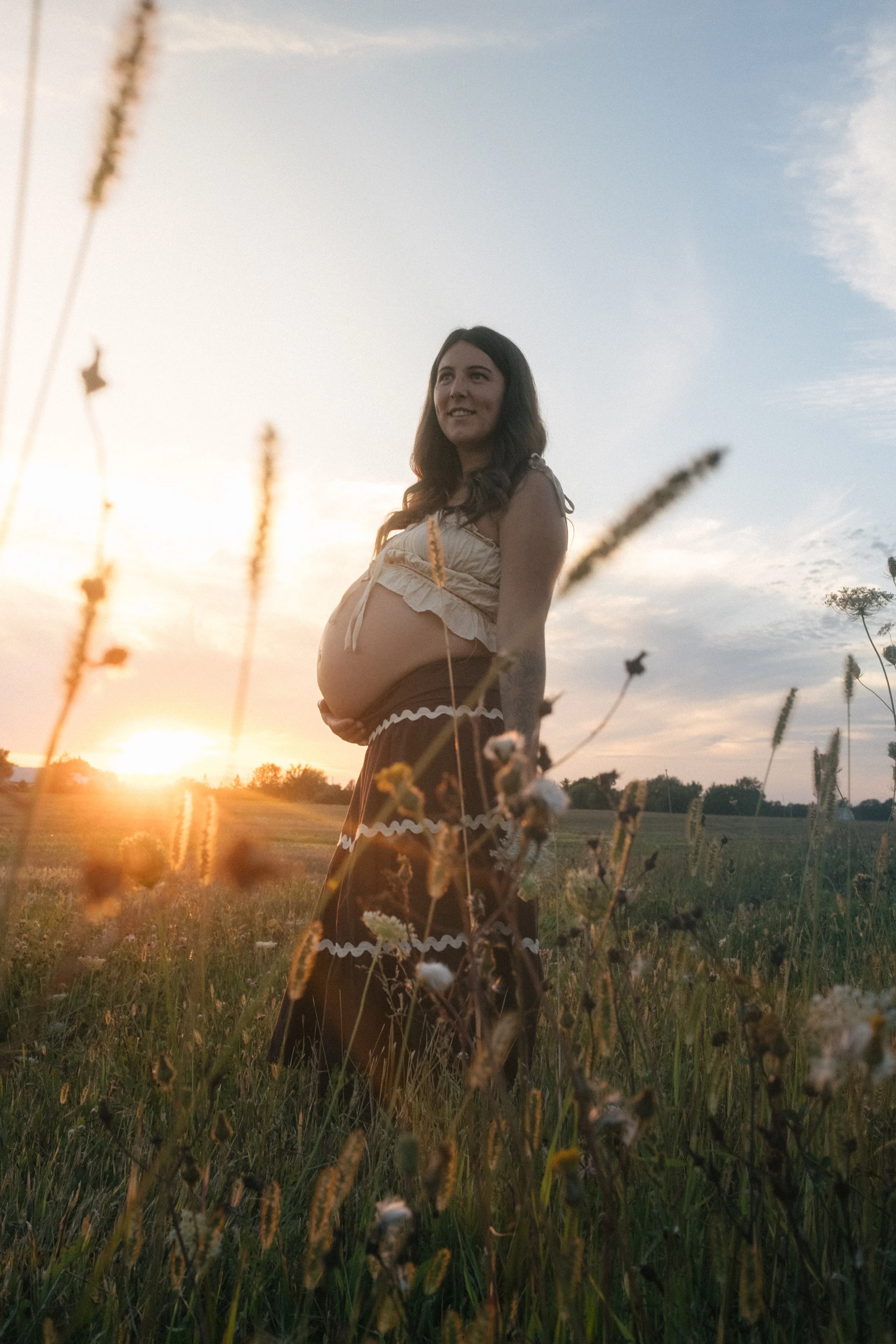 A pregnant woman standing in a grassy field during sunset, smiling and looking away from the camera.