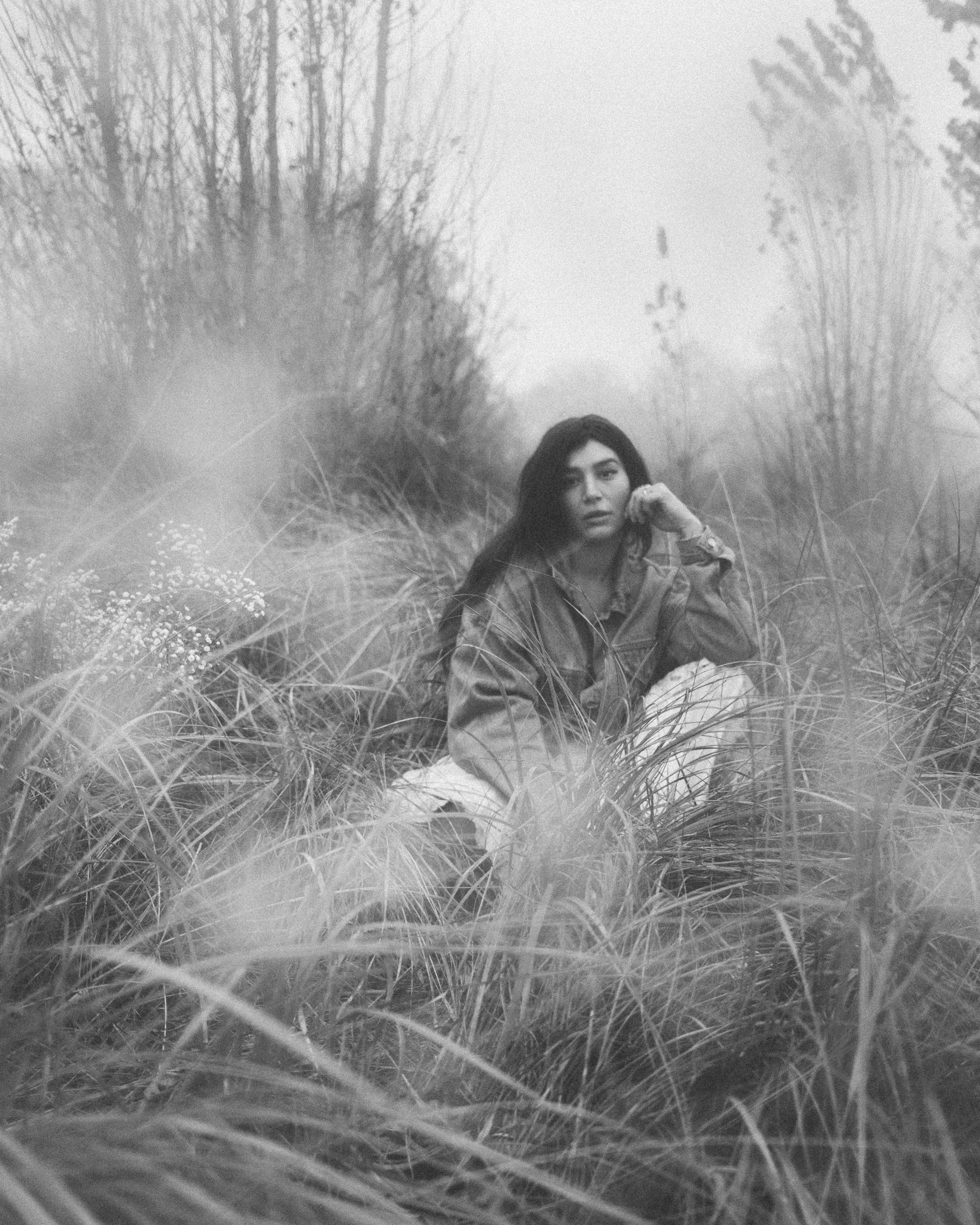 Black and white photo of a young woman sitting on grassy field surrounded by tall grass and trees, with a contemplative expression.