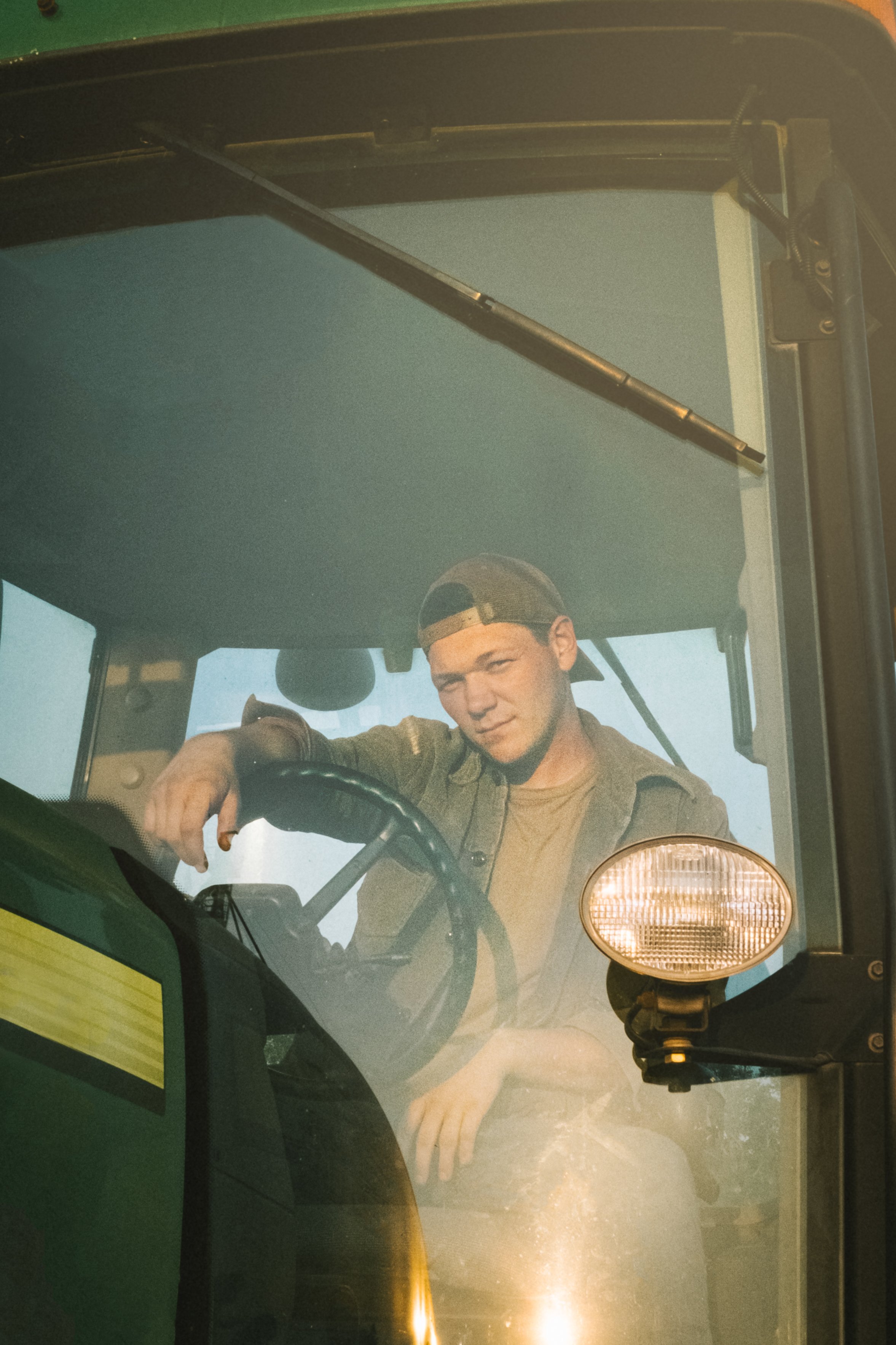 A young man sitting inside a tractor cab, wearing a baseball cap backwards, looking out through the glass window.