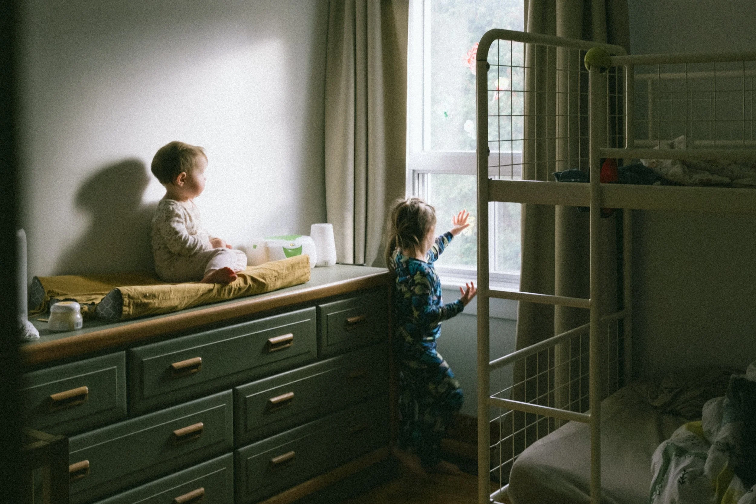 Indoor portrait of two young girls in a sunlit bedroom, one sitting on a dresser / diaper change table and the other standing by the window, gazing outside, with gentle natural light illuminating the scene