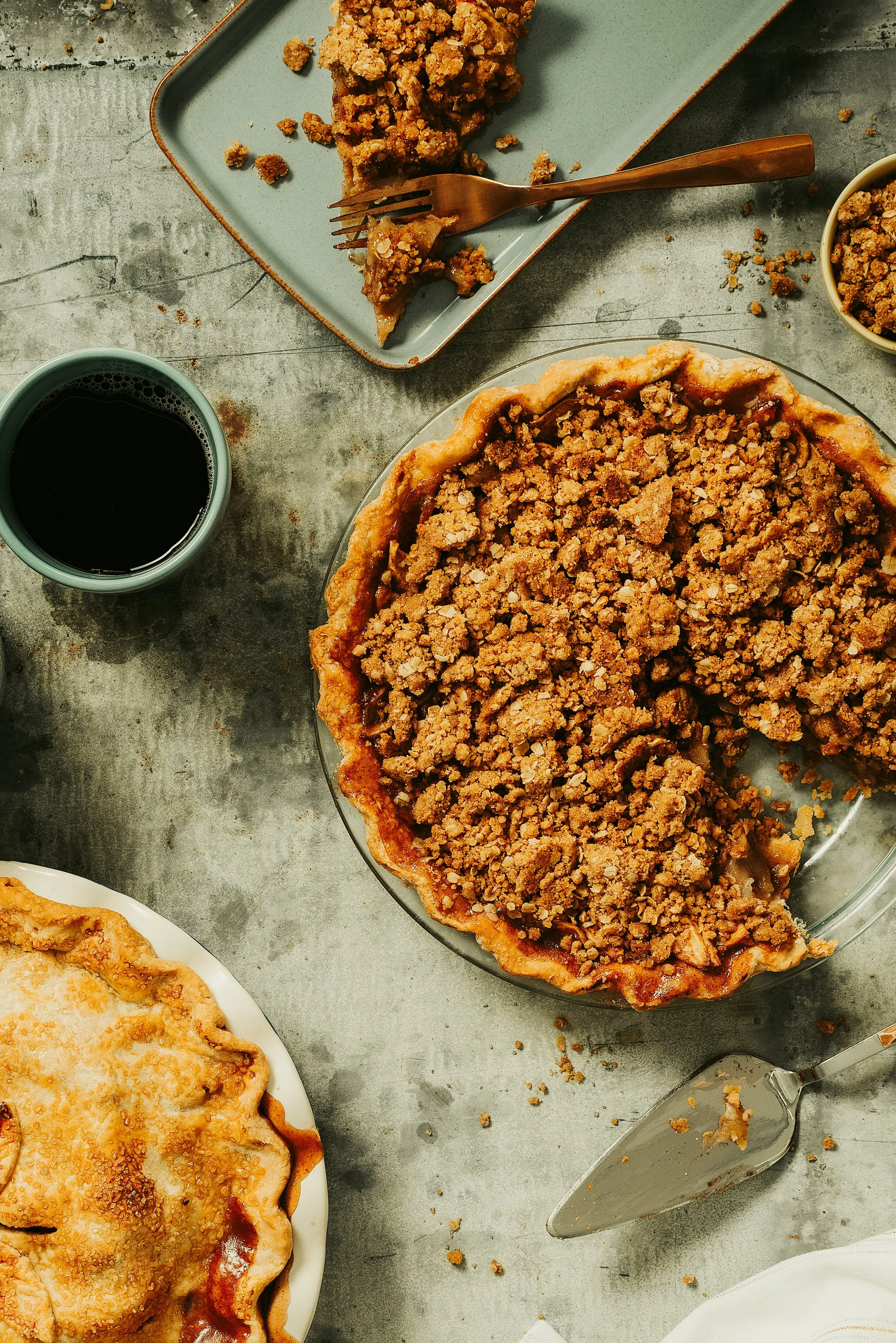 Slices of apple pie with crumb topping on a white plate, a partially eaten crumb-topped pie in a glass dish, a cup of coffee, a slice of pie on a teal plate with a fork, and small bowls of crumb topping on a gray textured surface.