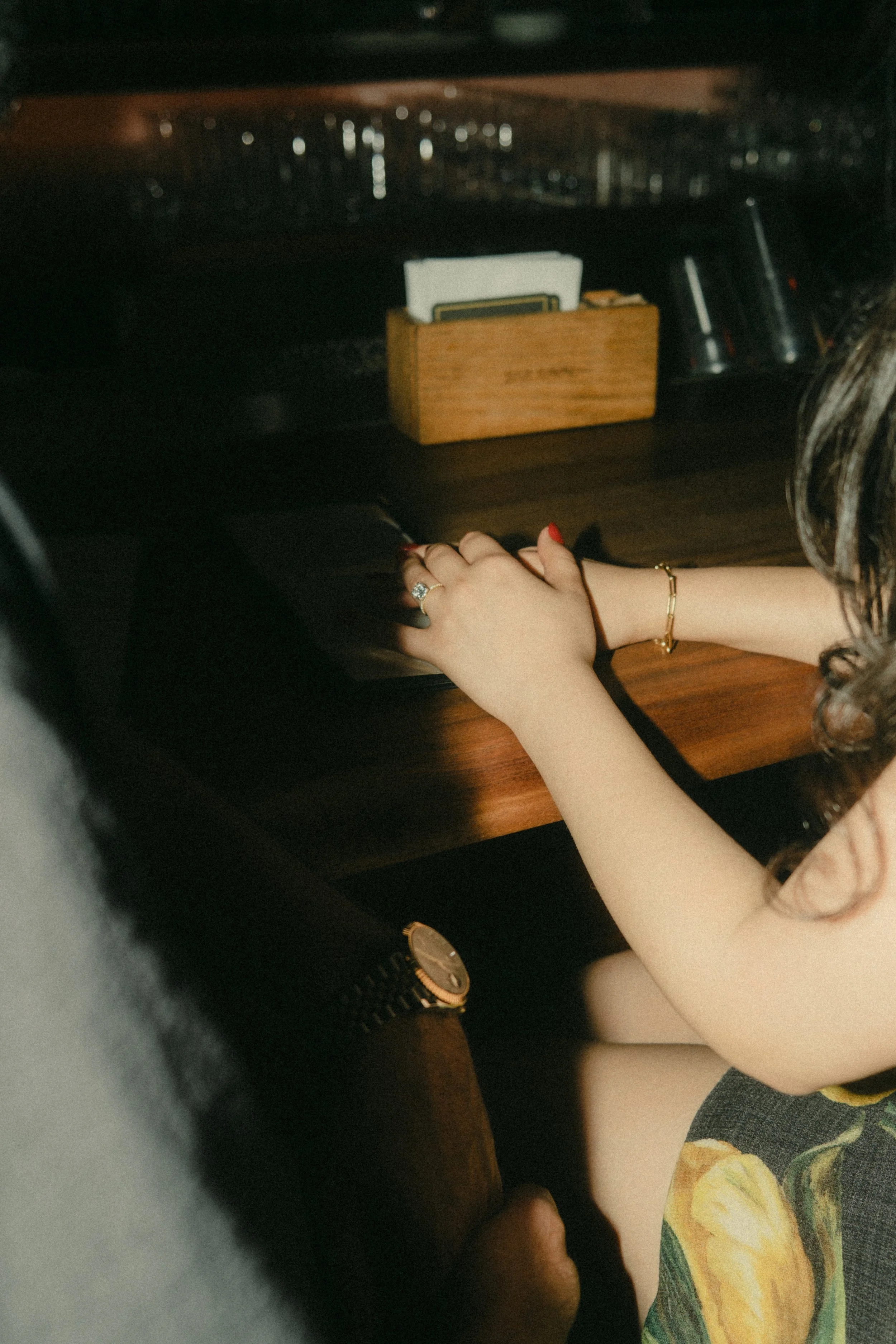 A woman with curly hair sitting at a wooden table, wearing a floral dress, a gold bracelet, and a watch, with her hands resting on the table and a ring on her finger.