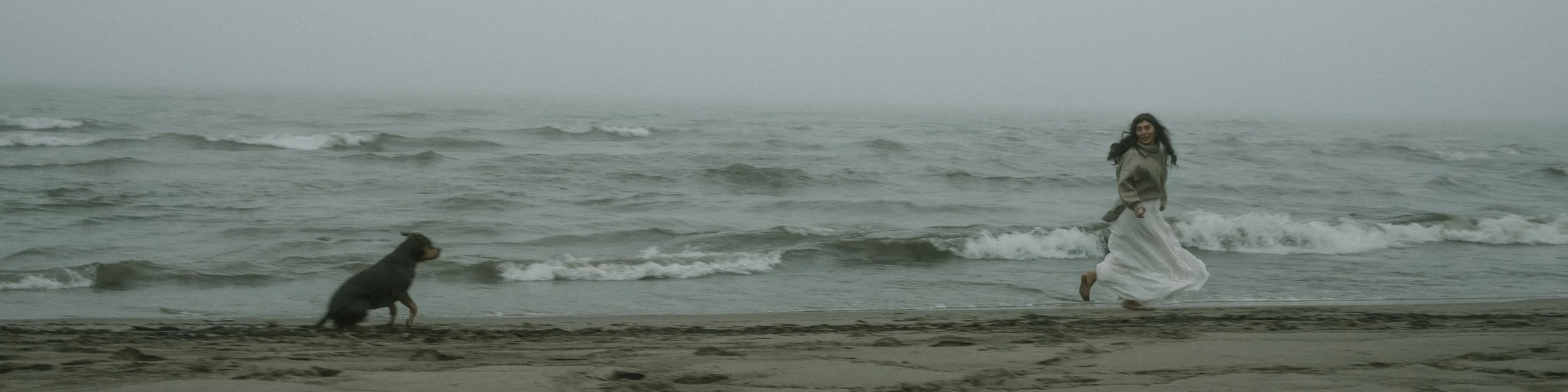 A woman wearing a gray sweater and long white skirt running along the beach with a dog near the shoreline on a foggy day.