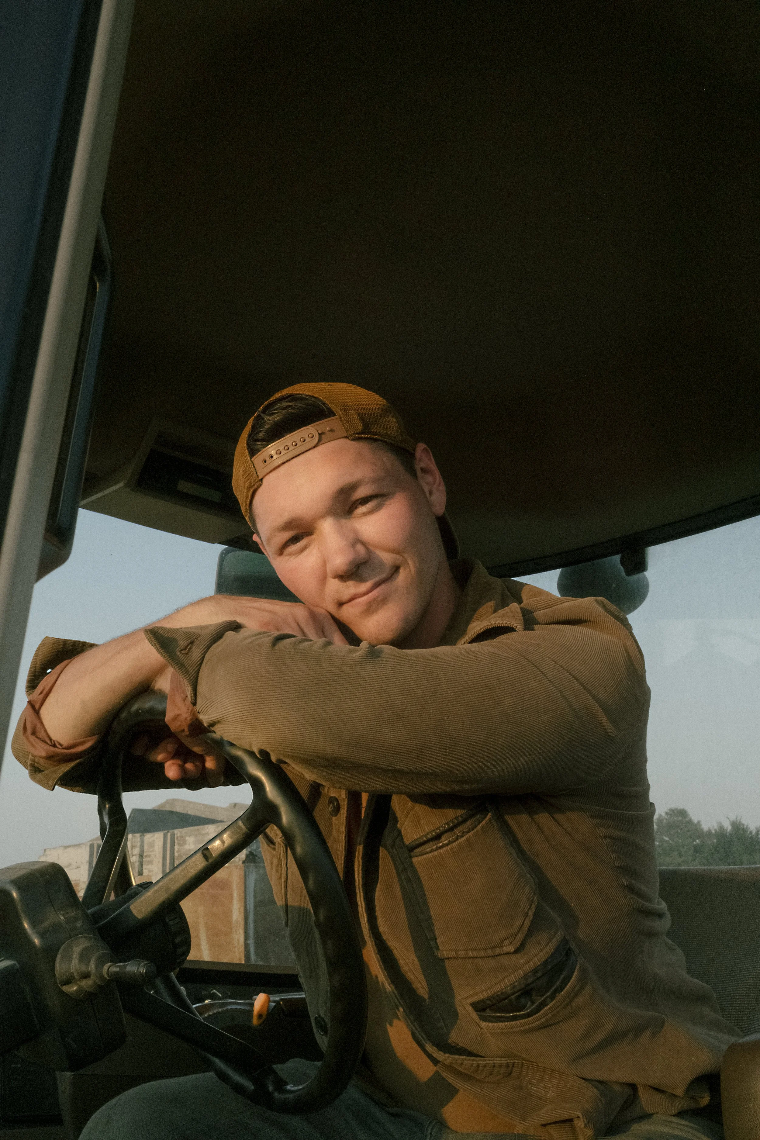 A young man in a brown jacket and backwards cap leaning on a tractor's steering wheel, smiling at the camera.