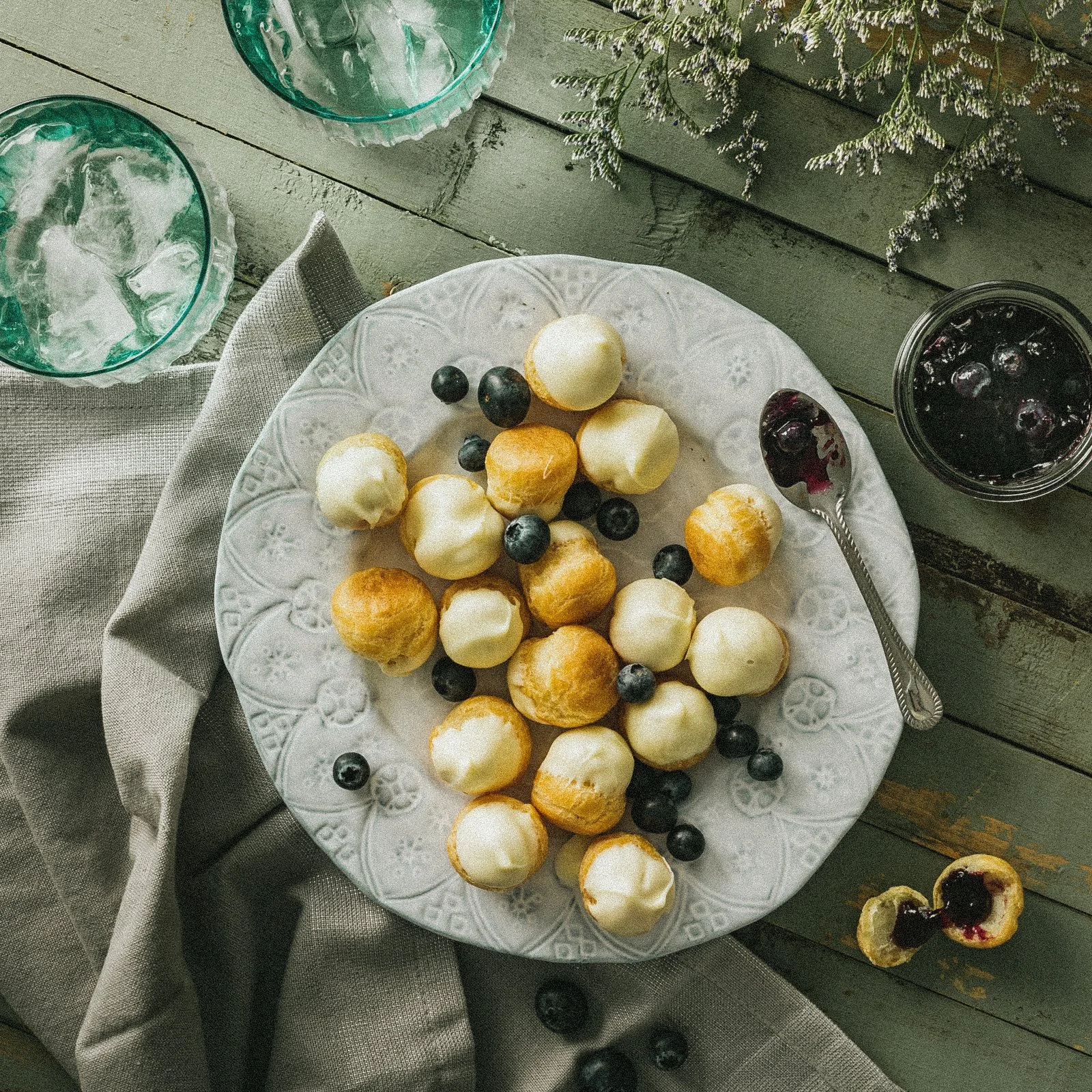 A white decorative plate with assorted small cream-filled and golden-brown pastries, topped with blueberries, resting on a rustic wooden table with a beige napkin nearby, with glasses of ice water and a jar of blueberries with syrup around.