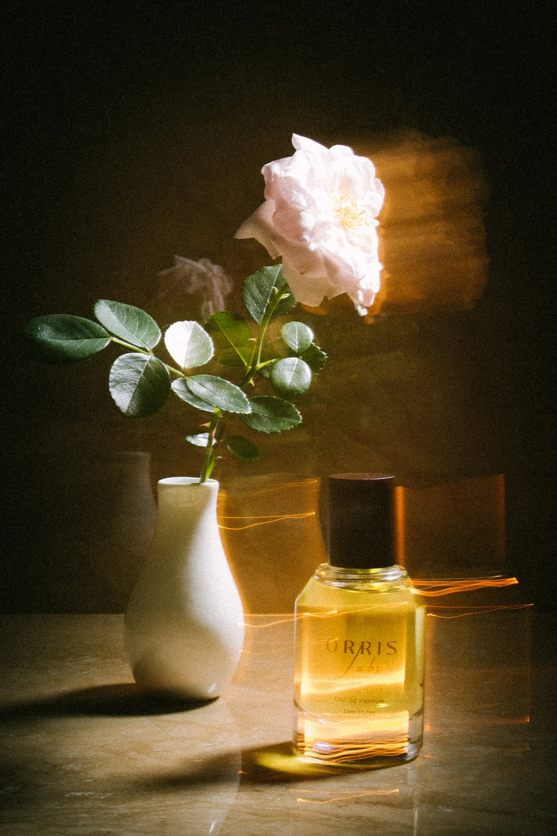A small white vase with a pink flower and green leaves, next to a bottle of Orris perfume on a wooden surface with lighting effects.