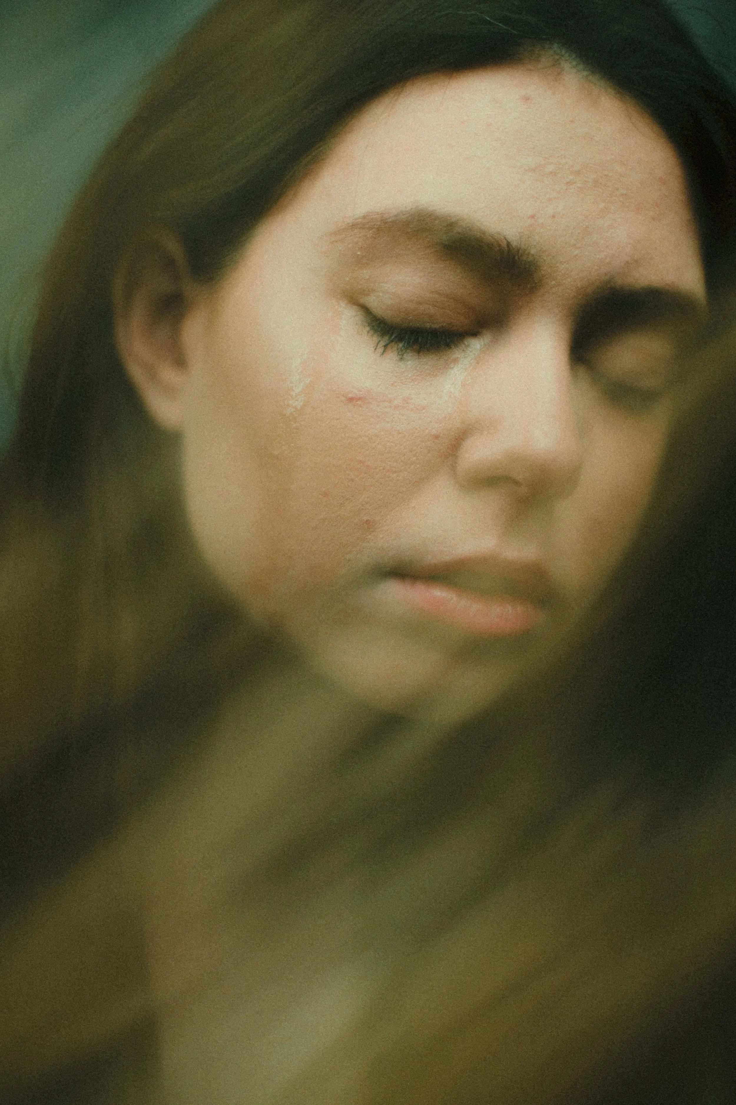 Close-up of a woman with closed eyes and long dark hair. She has some white marks or scars on her cheeks.