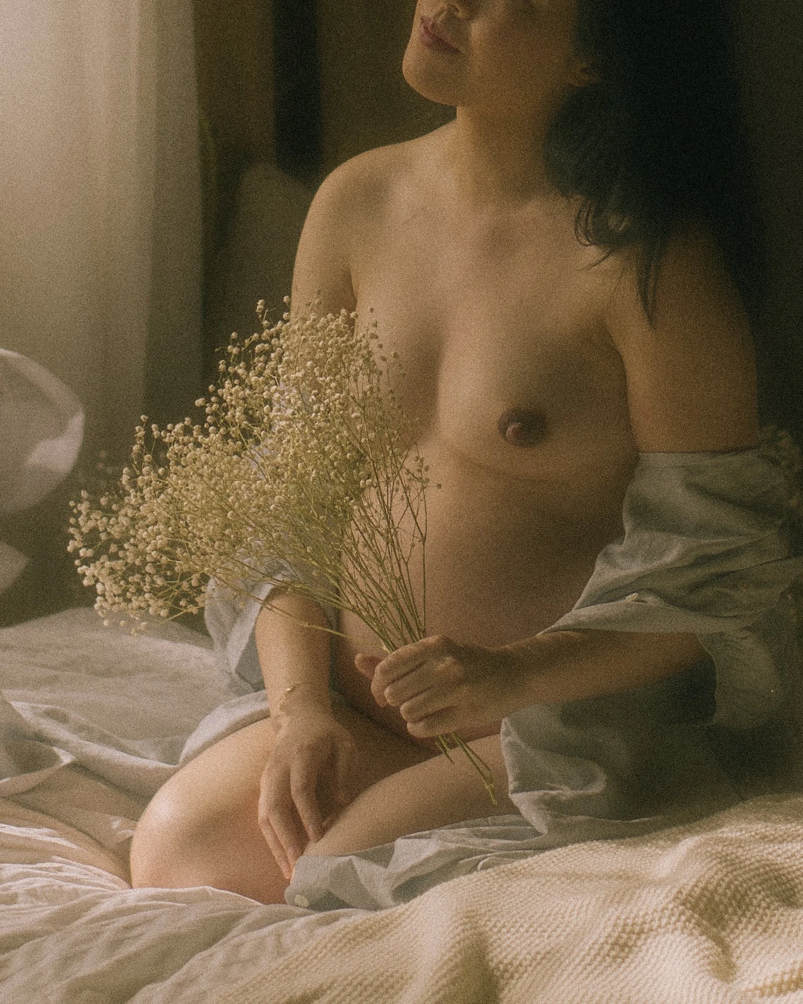 A woman sitting on a bed holding a bouquet of dried flowers, partially covered by a sheet, with a warm, soft lighting.