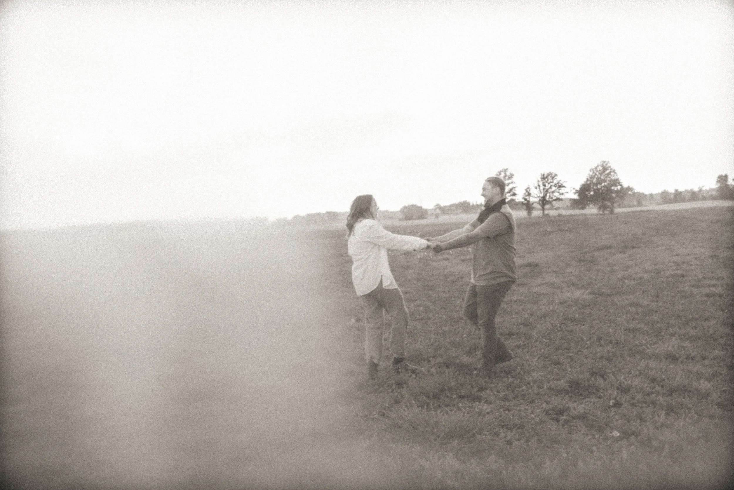 Dreamy black and white photograph of a couple holding hands and spinning in an open field with sparse trees, soft white light streaming from one side, with gentle blur and lens flare creating an ethereal, romantic atmosphere