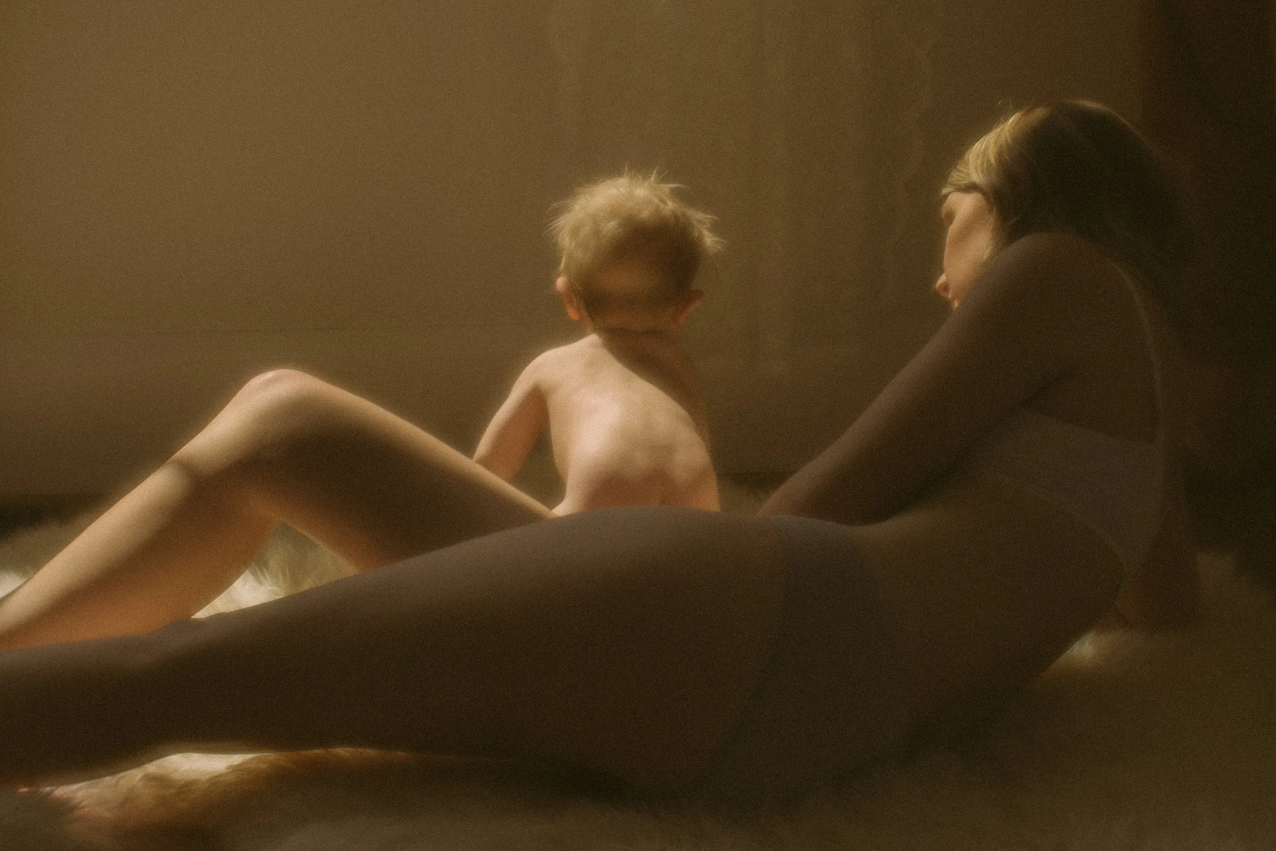 A woman and a child sitting on a bed in a dimly lit room, facing each other.