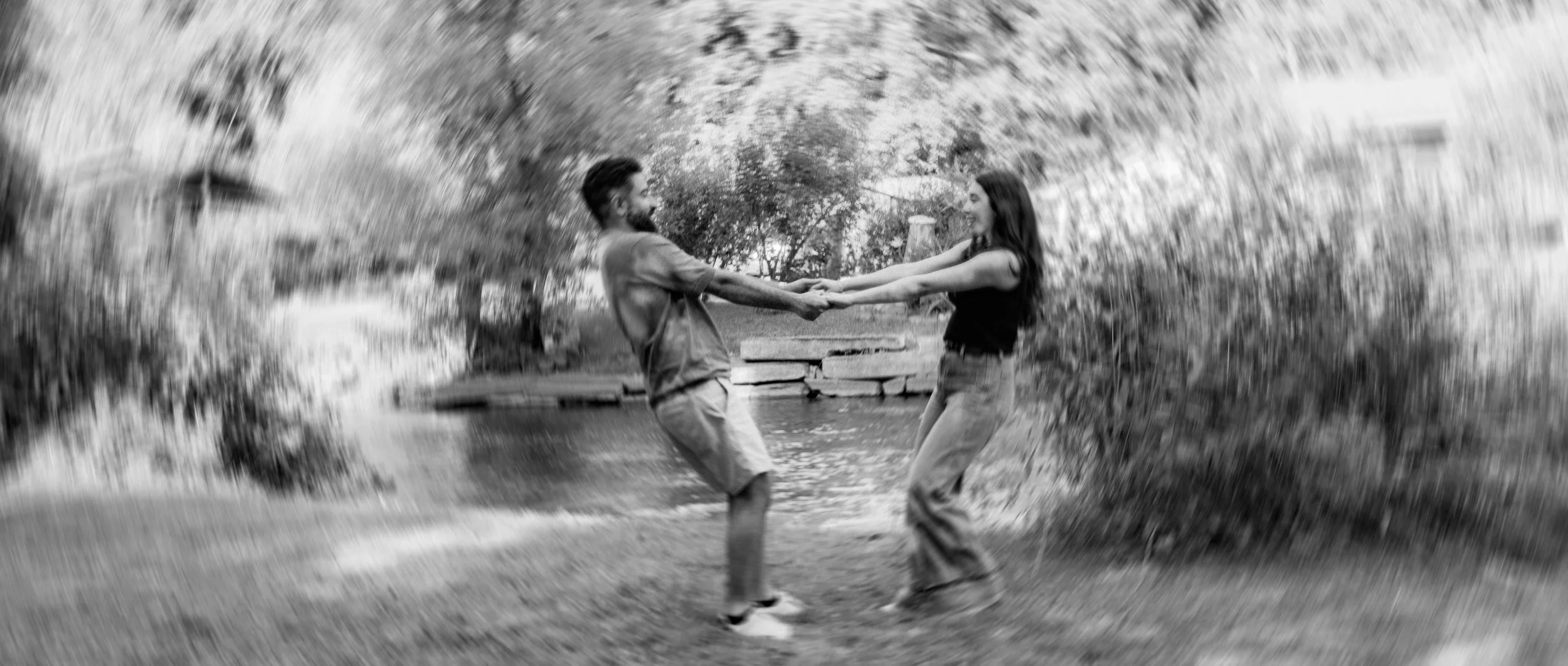 A black and white photo of a man and woman holding hands and smiling at each other while standing in a shallow creek, surrounded by trees and natural scenery.