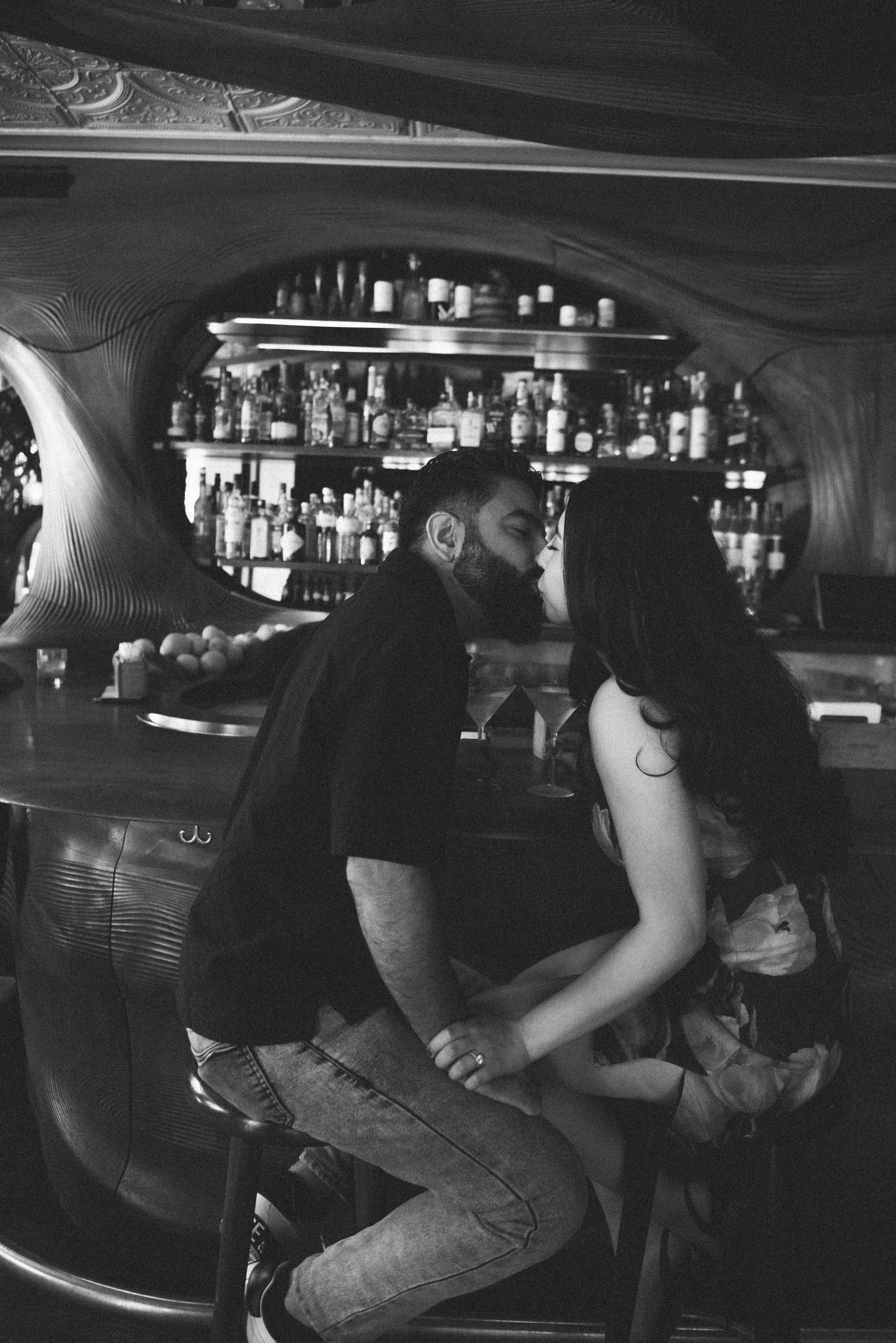A black and white photo of a man and woman sitting close together at a bar, about to kiss, with a backdrop of shelves filled with bottles of alcohol.