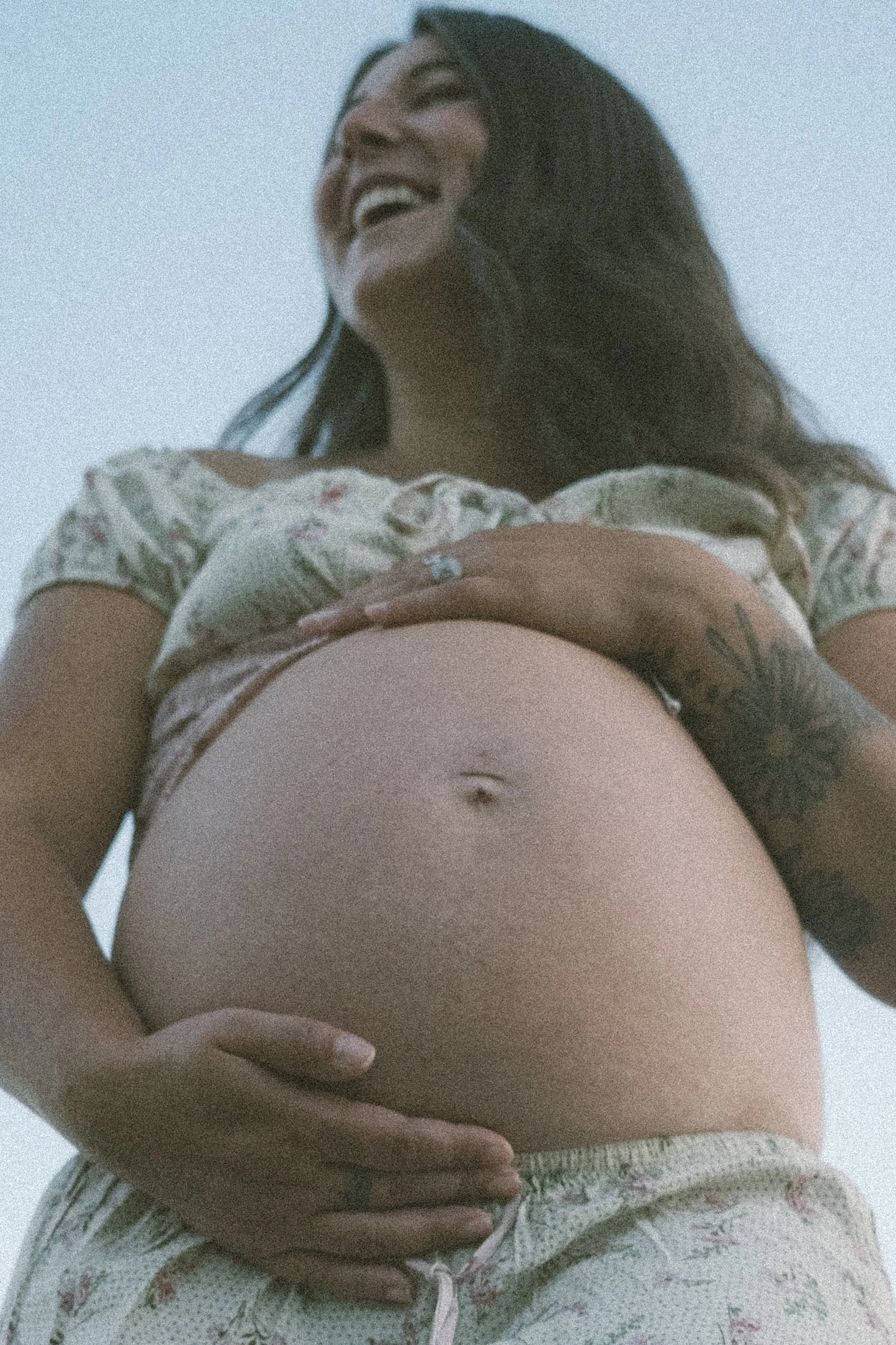 A woman is smiling and looking up, lifting her shirt to reveal her pregnant belly, with her hand resting on it.