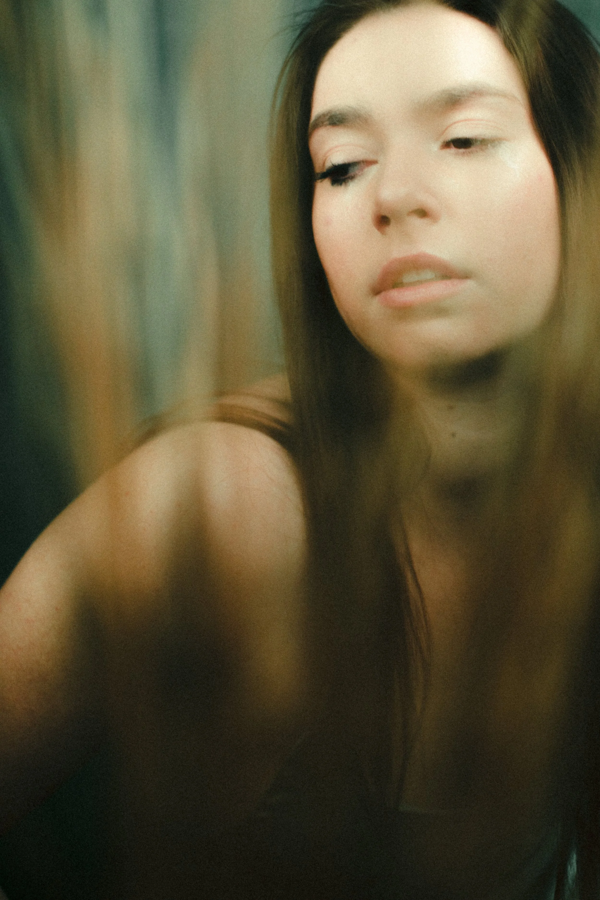 Close-up of a young woman with long brown hair, fair skin, and makeup, with a contemplative expression, looking slightly downward, in soft lighting.