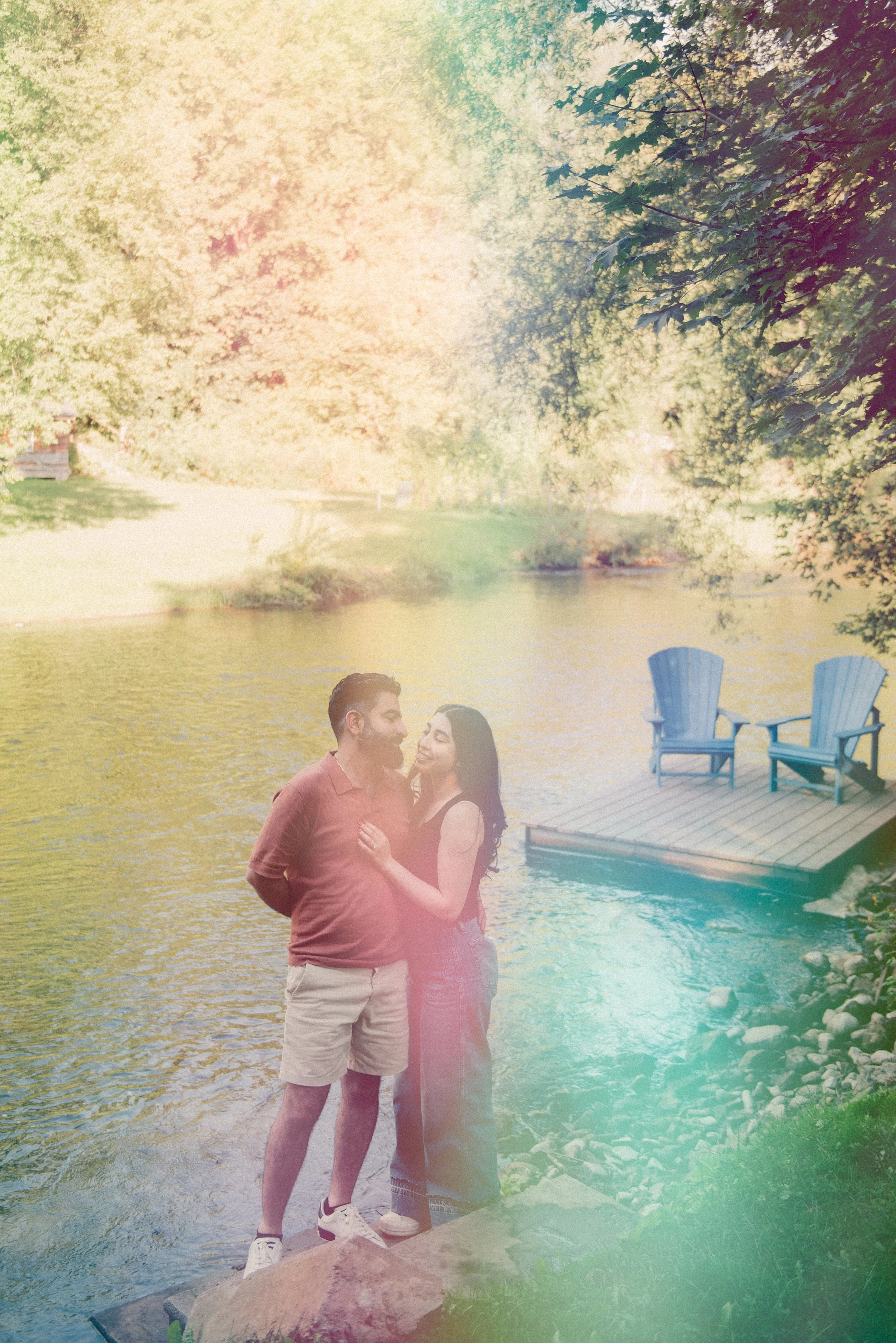 A couple standing by a lake with two Adirondack chairs on a wooden dock, surrounded by green trees, with sunlight filtering through the leaves.