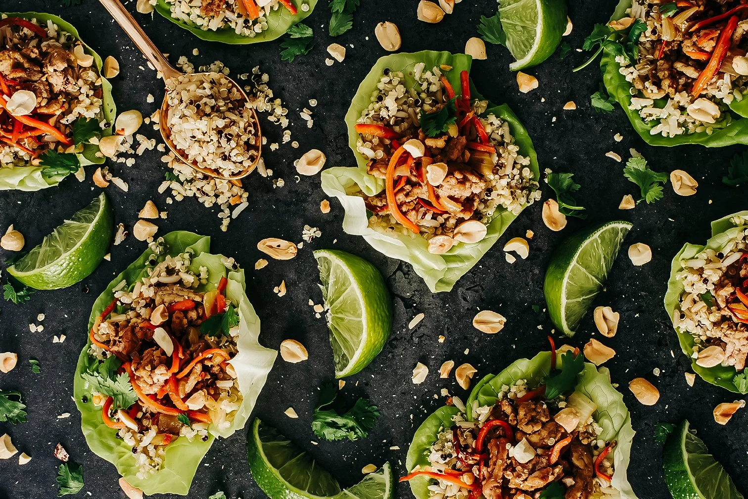 Lettuce wraps filled with ground meat, vegetables, and chopped peanuts, garnished with cilantro, accompanied by lime wedges, all placed on a dark surface.