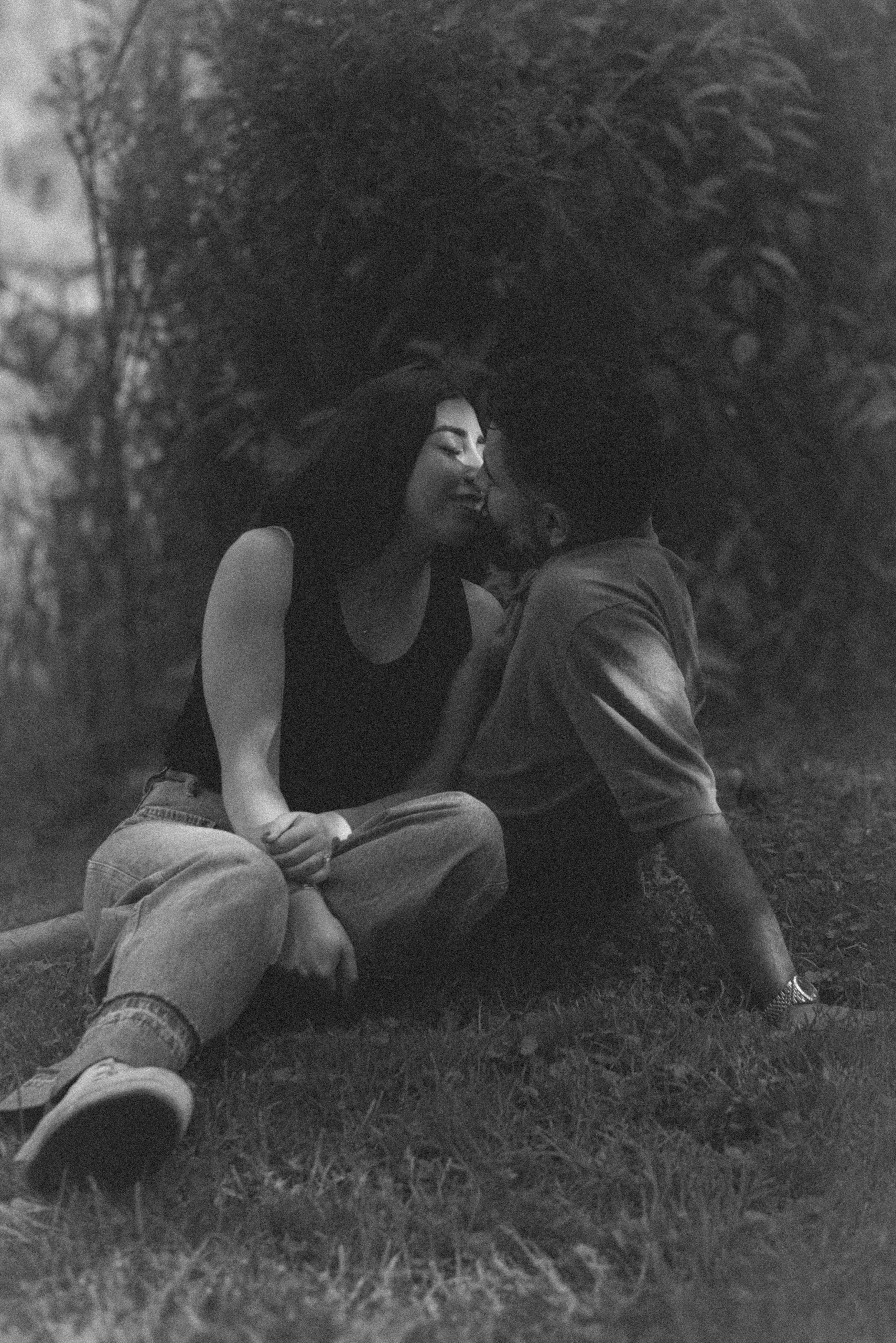 Black and white outdoor portrait of a young couple about to share a kiss while sitting on the grass, surrounded by trees, capturing a tender, intimate moment