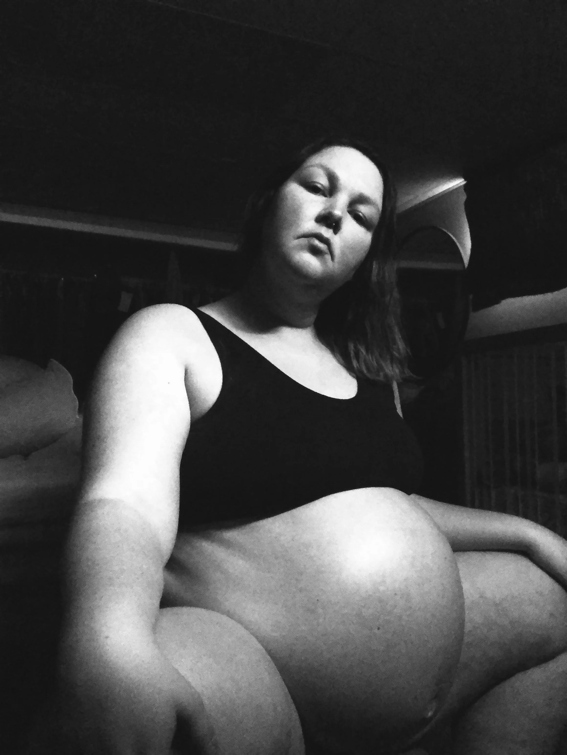 High-contrast black and white self-portrait of a very pregnant woman during labor, sitting with legs bent in a dark room, wearing a black sports bra, with shoulder-length hair and a strong, focused expression, capturing the intensity and intimacy 