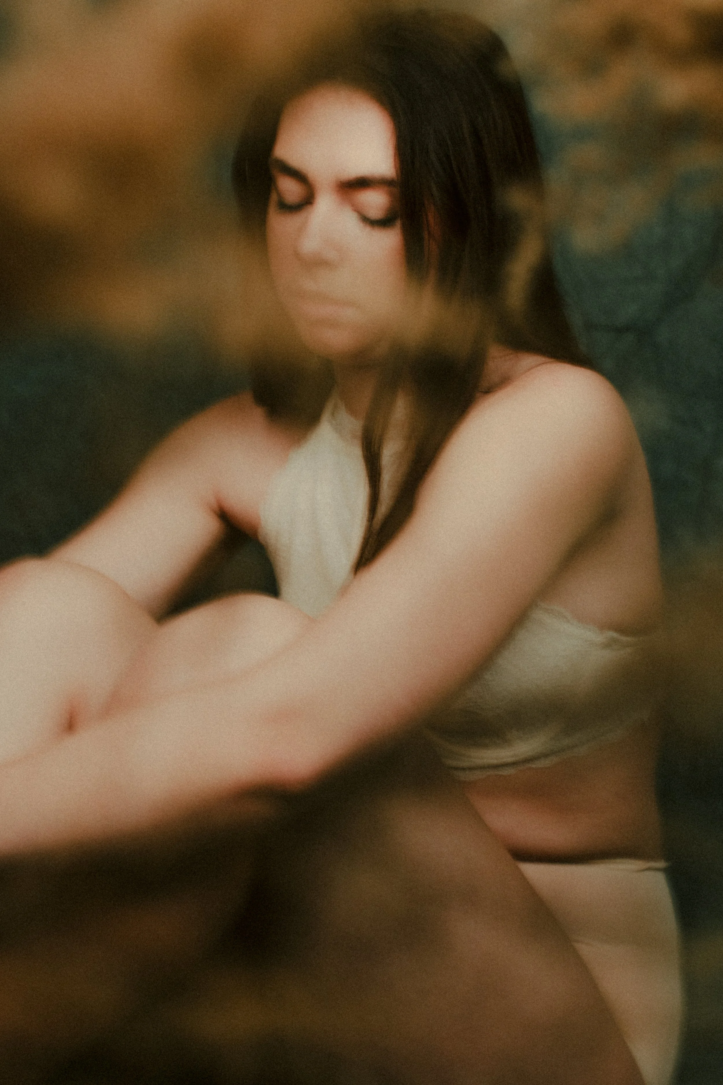 A woman with dark hair and pale skin, closed eyes, wearing a sleeveless beige top, sitting with her arms crossed and a calm expression.