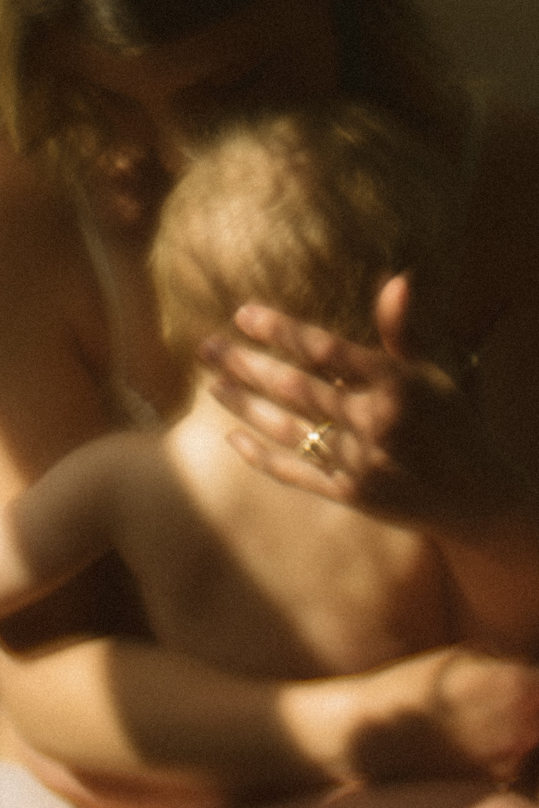 A woman with short hair covering her face with her hand, wearing a ring, and embracing back of baby's head.