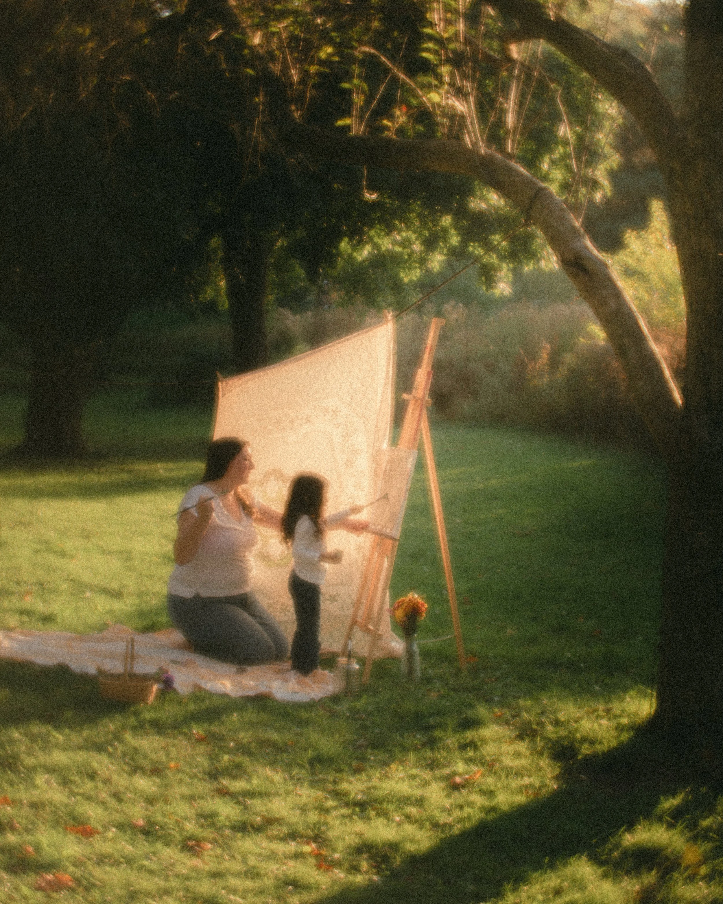 A woman and a girl painting outdoors on a sunny day, kneeling on a blanket with an easel and canvas set up under trees.