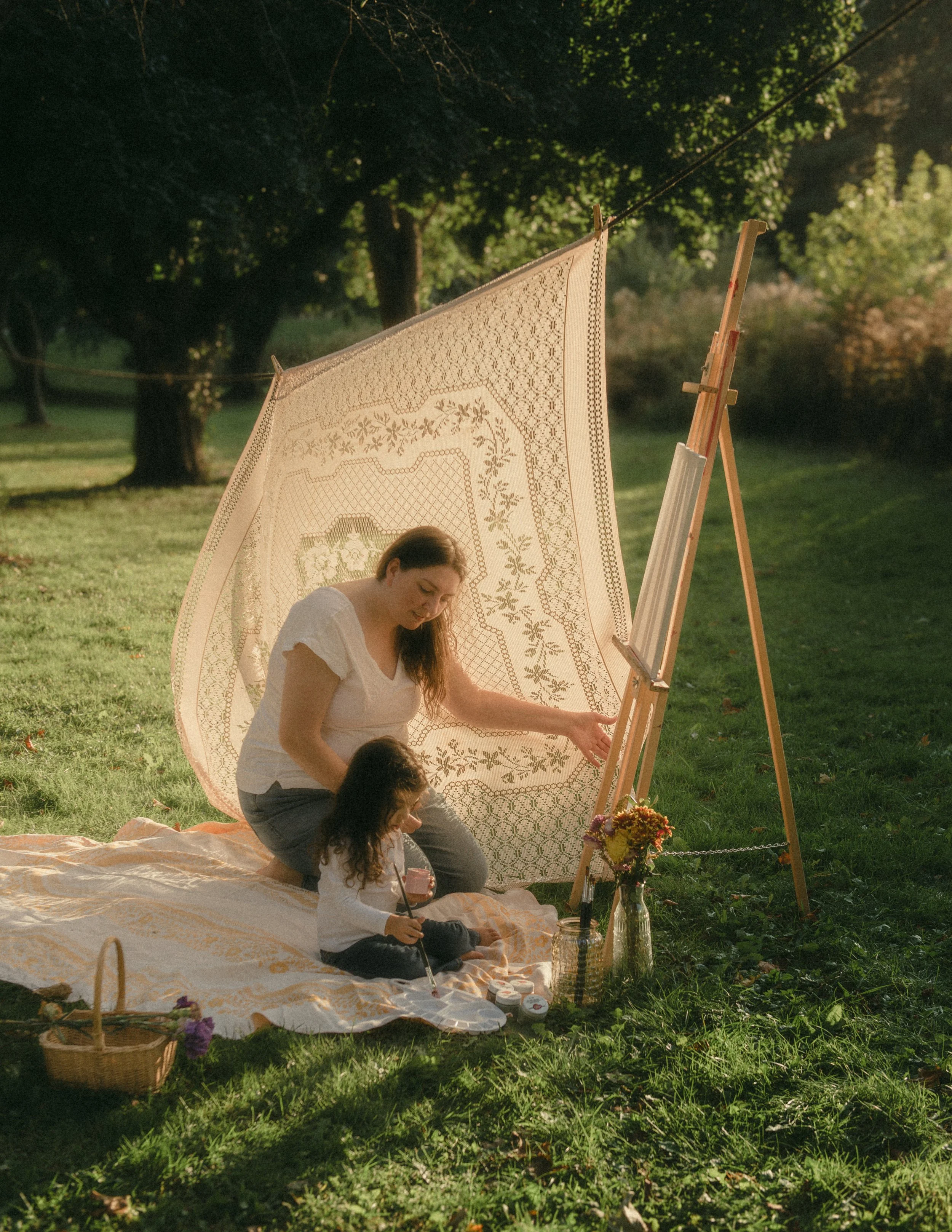 A woman and a young girl are sitting on a blanket outdoors, painting on large canvases set up on easels under a decorated canopy, with trees and grassy area in the background.