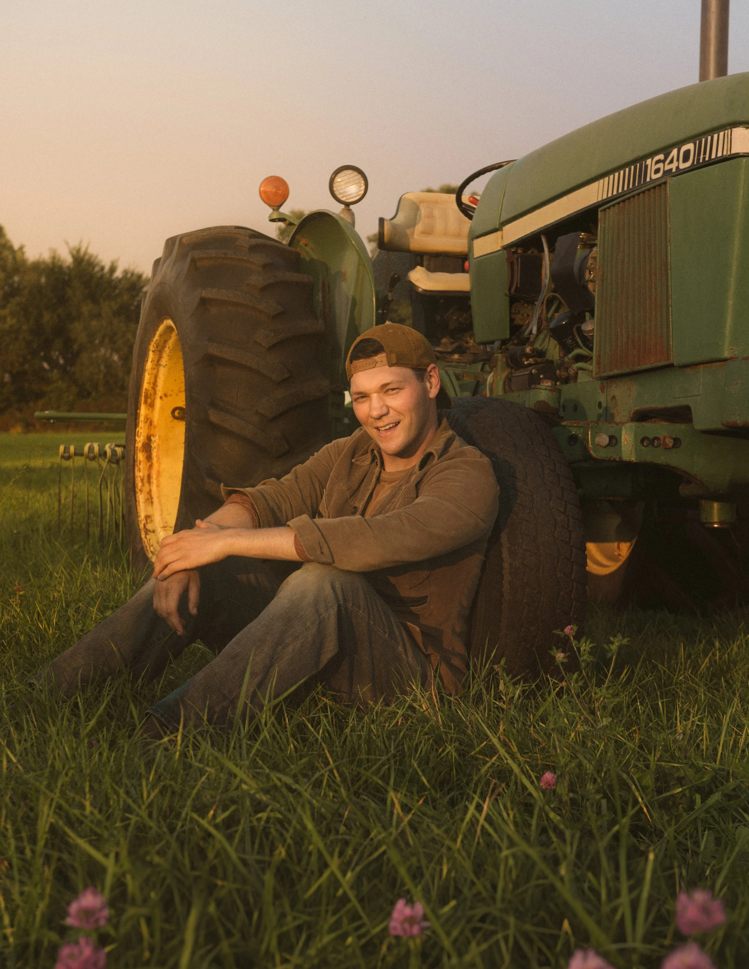 A young man sitting on grass in front of a tractor, smiling at the camera during sunset.