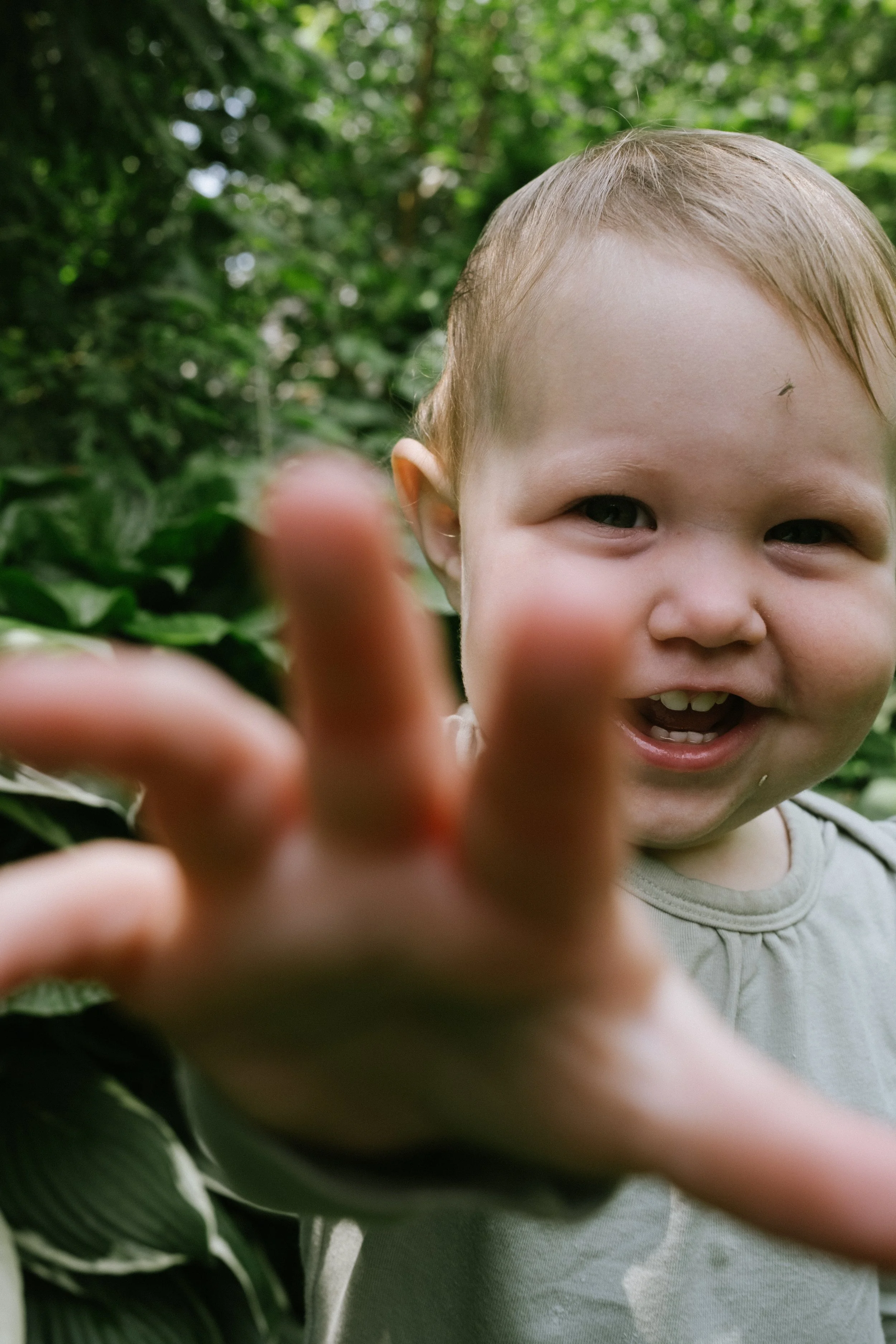 Young child smiling and reaching out with hand in front of green foliage background