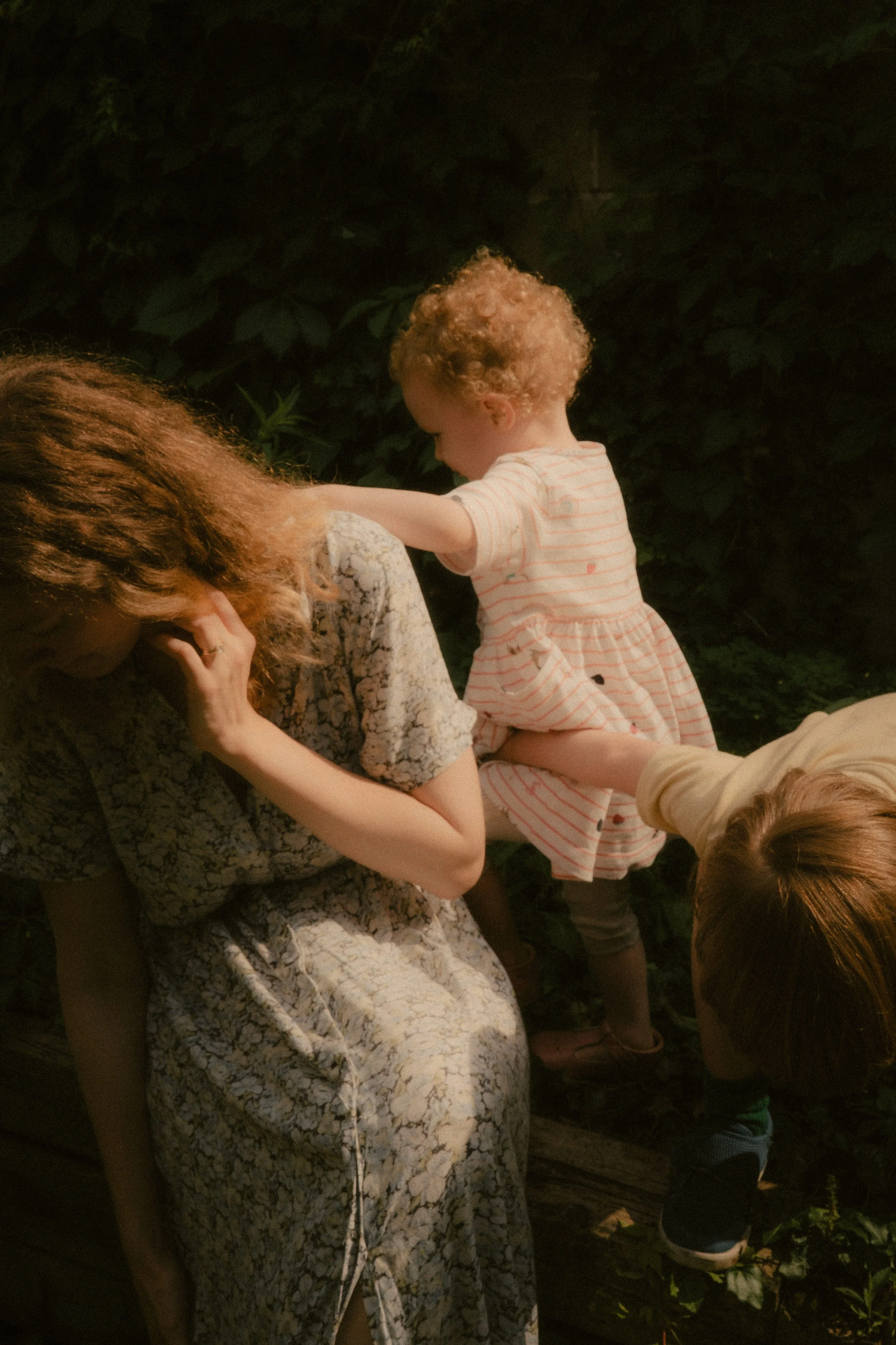 A woman sitting on a wooden bench outdoors, with a young girl in a striped dress touching her shoulder, and another child leaning in to her side, all surrounded by greenery.