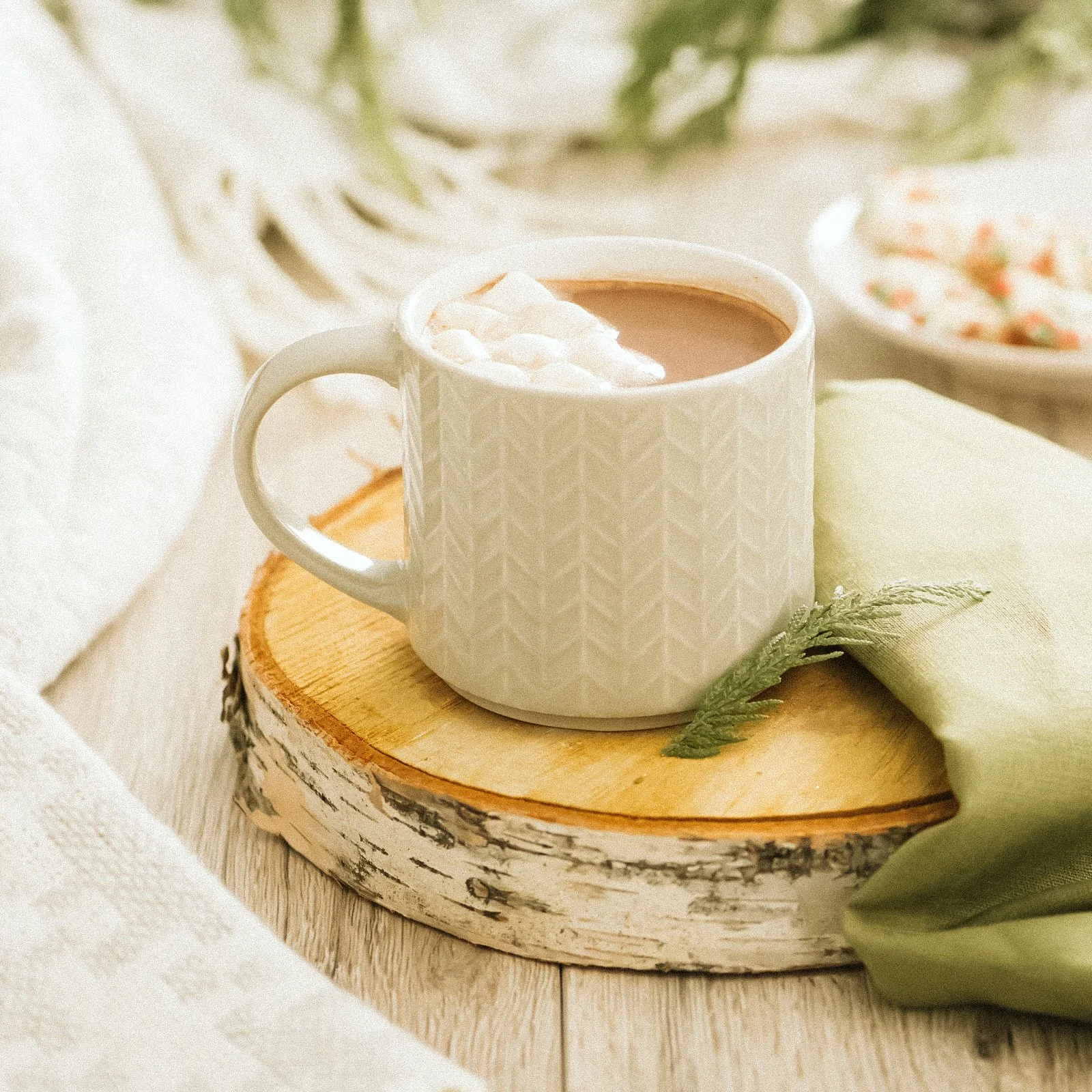 A white mug with a textured pattern, filled with hot chocolate topped with marshmallows, sitting on a log slice. There is a light green cloth and a fern leaf beside it, with a blurred bowl of cereal or dessert in the background.