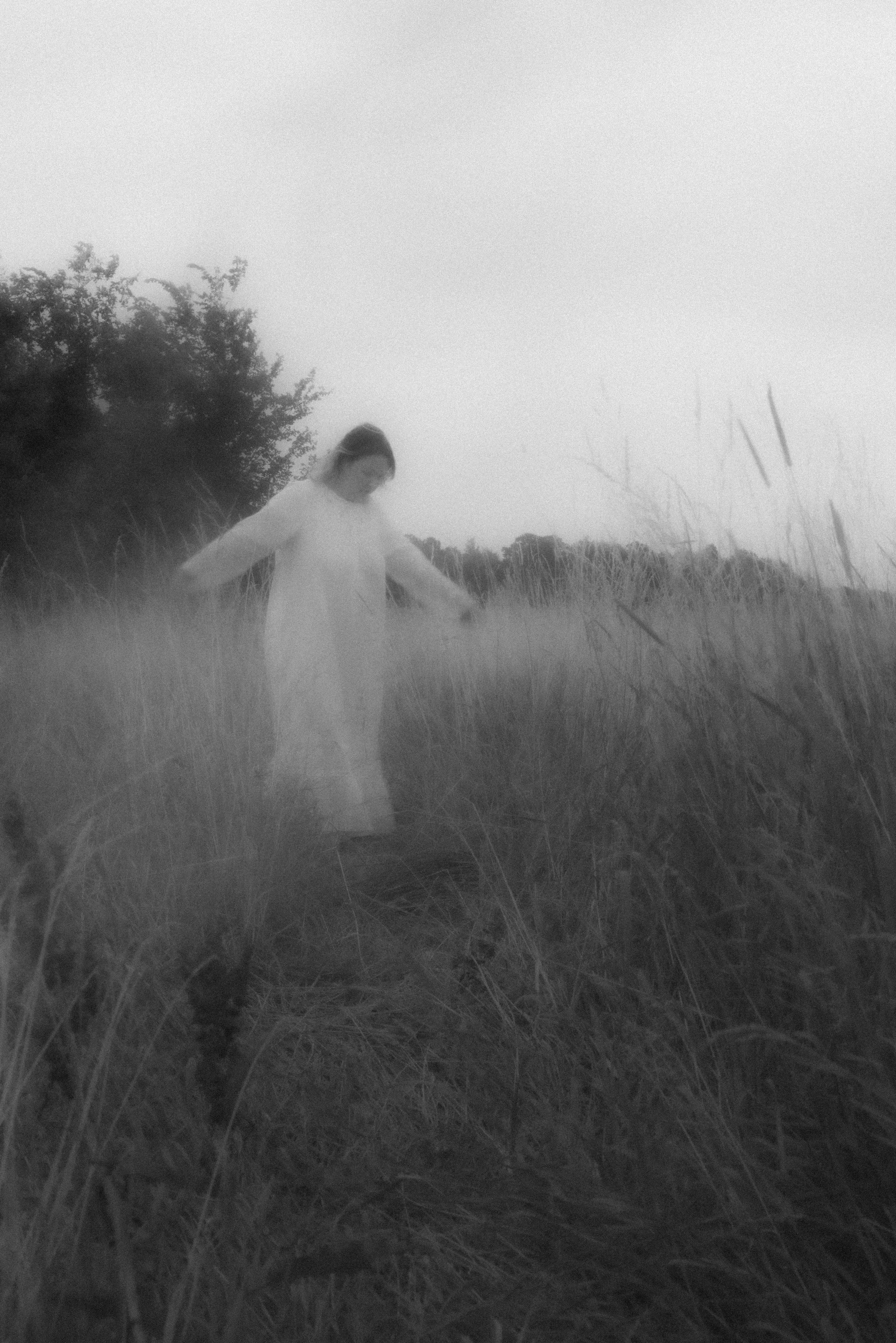 Artistic black and white portrait of a woman in a white nightgown walking through tall grass, captured with motion blur and a foggy, ethereal atmosphere, with trees in the background