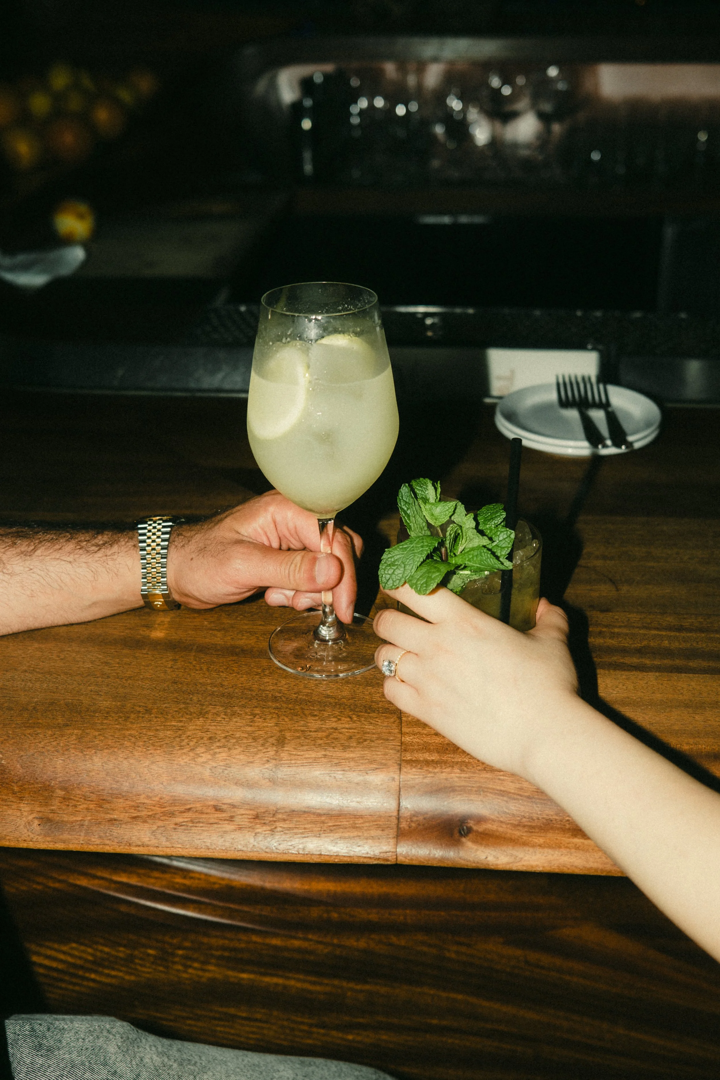 A person with a wristwatch holding a glass of lemonade with lemon slices, and another person with a ring holding a mojito garnished with mint leaves on a wooden table.