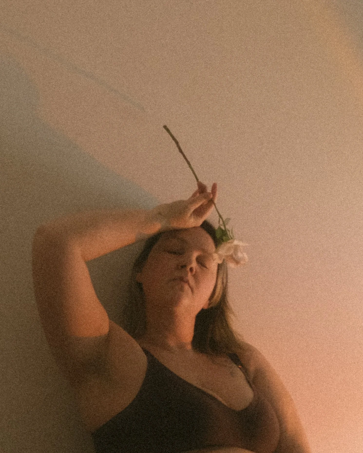 Artistic self-portrait of a woman with closed eyes, holding a rose draping over her face, bathed in warm pink and orange light resembling a glowing lamp, creating a soft, intimate atmosphere