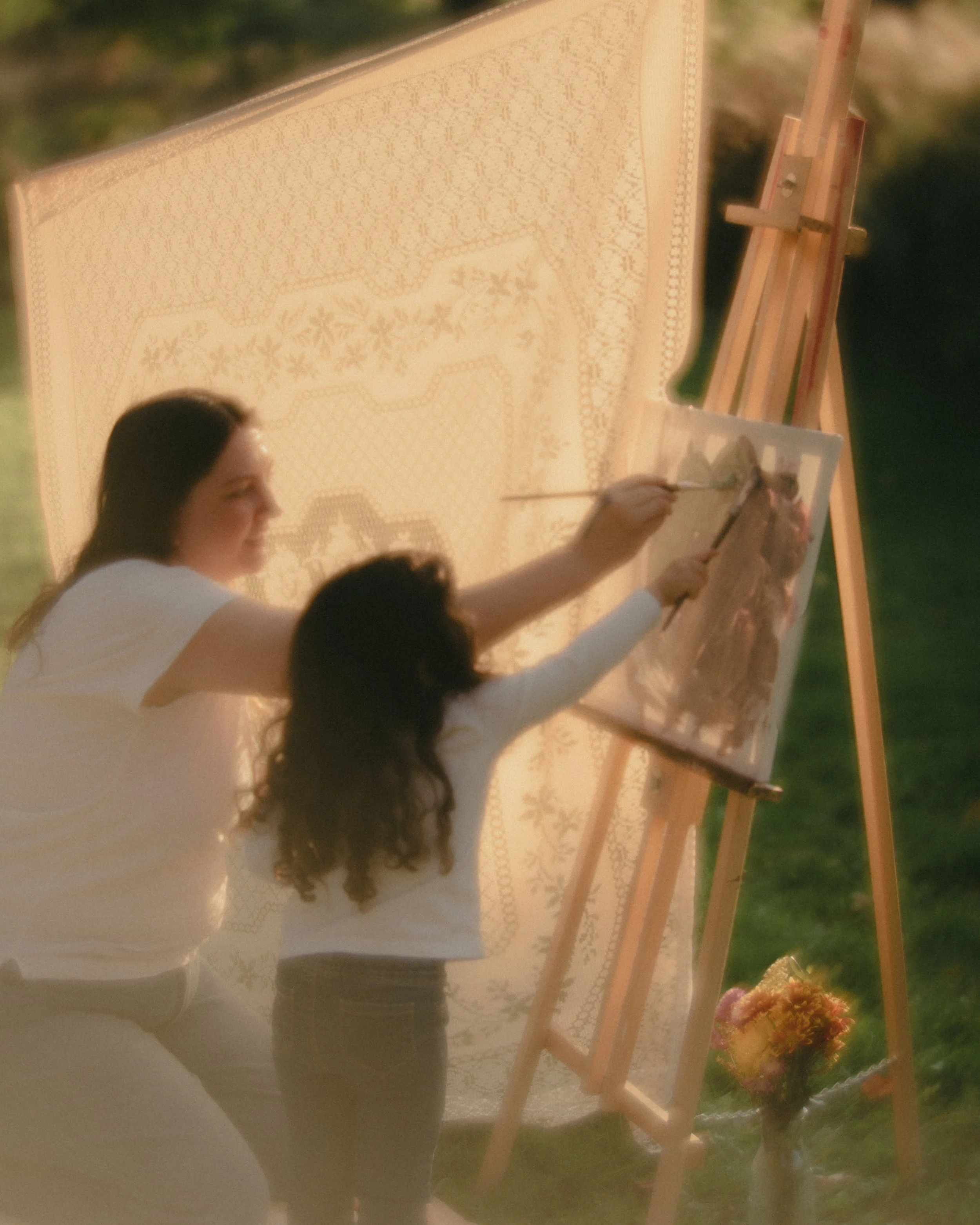 A woman and a young girl painting on a canvas outdoors during sunset, with a decorative screen and a bouquet of flowers nearby.