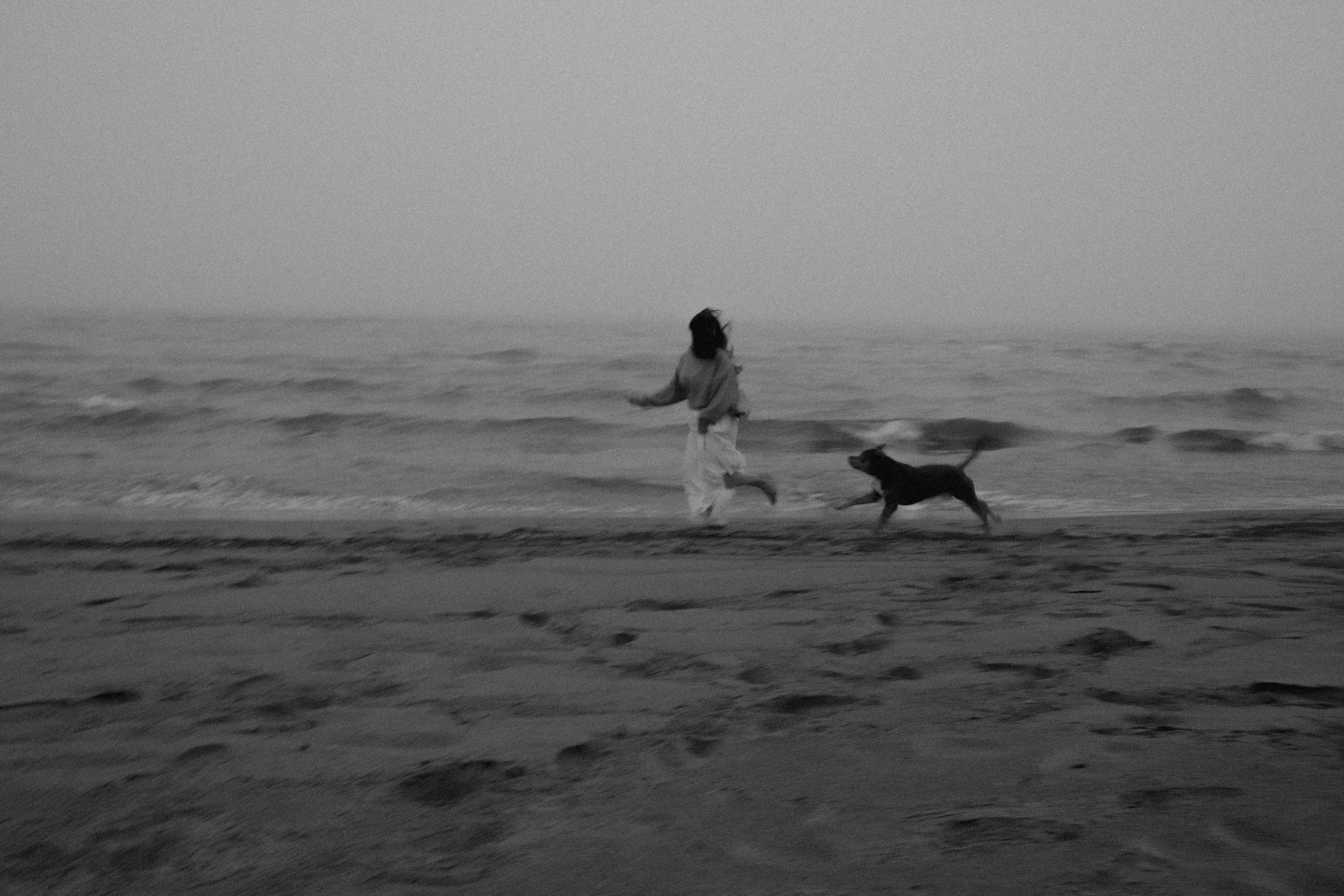 A person running along the shoreline with a dog on the beach, waves in the background, black and white photo.