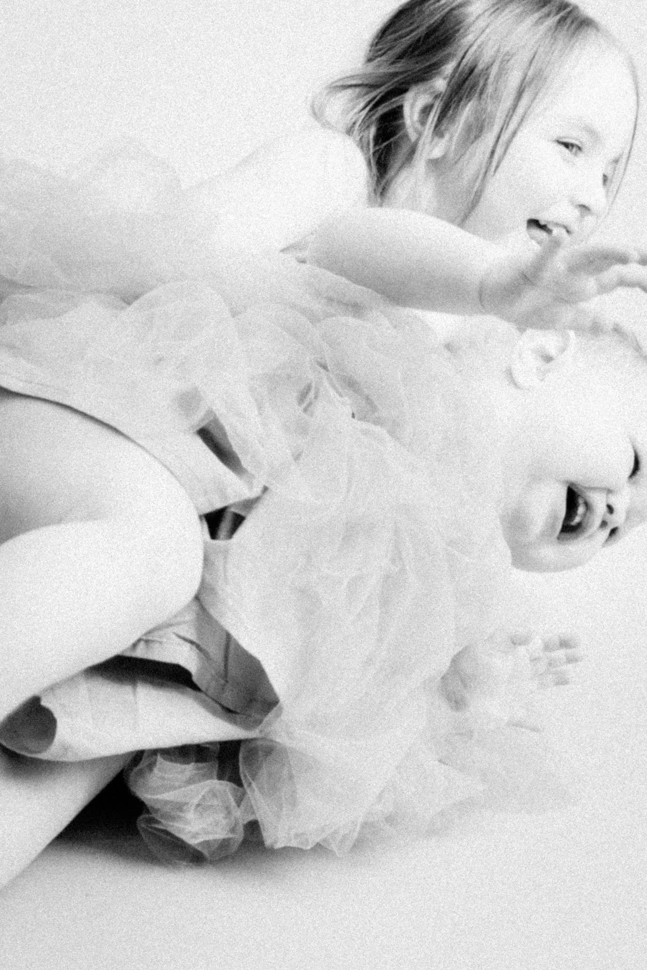 Dynamic black and white portrait of a young girl in a tutu dress lying on the floor with arms outstretched, holding a toddler who is screaming, capturing a playful, joyful, and chaotic moment