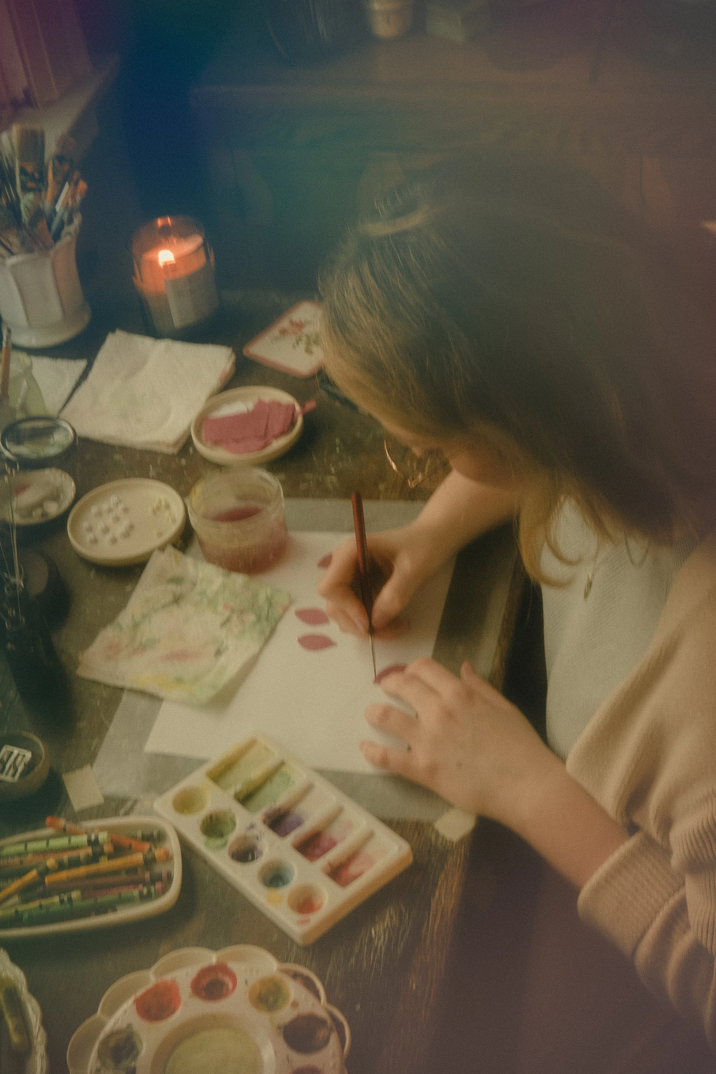 A person seated at a table, painting on a piece of paper with watercolor paints. The table is filled with paint palettes, brushes, and various art supplies.