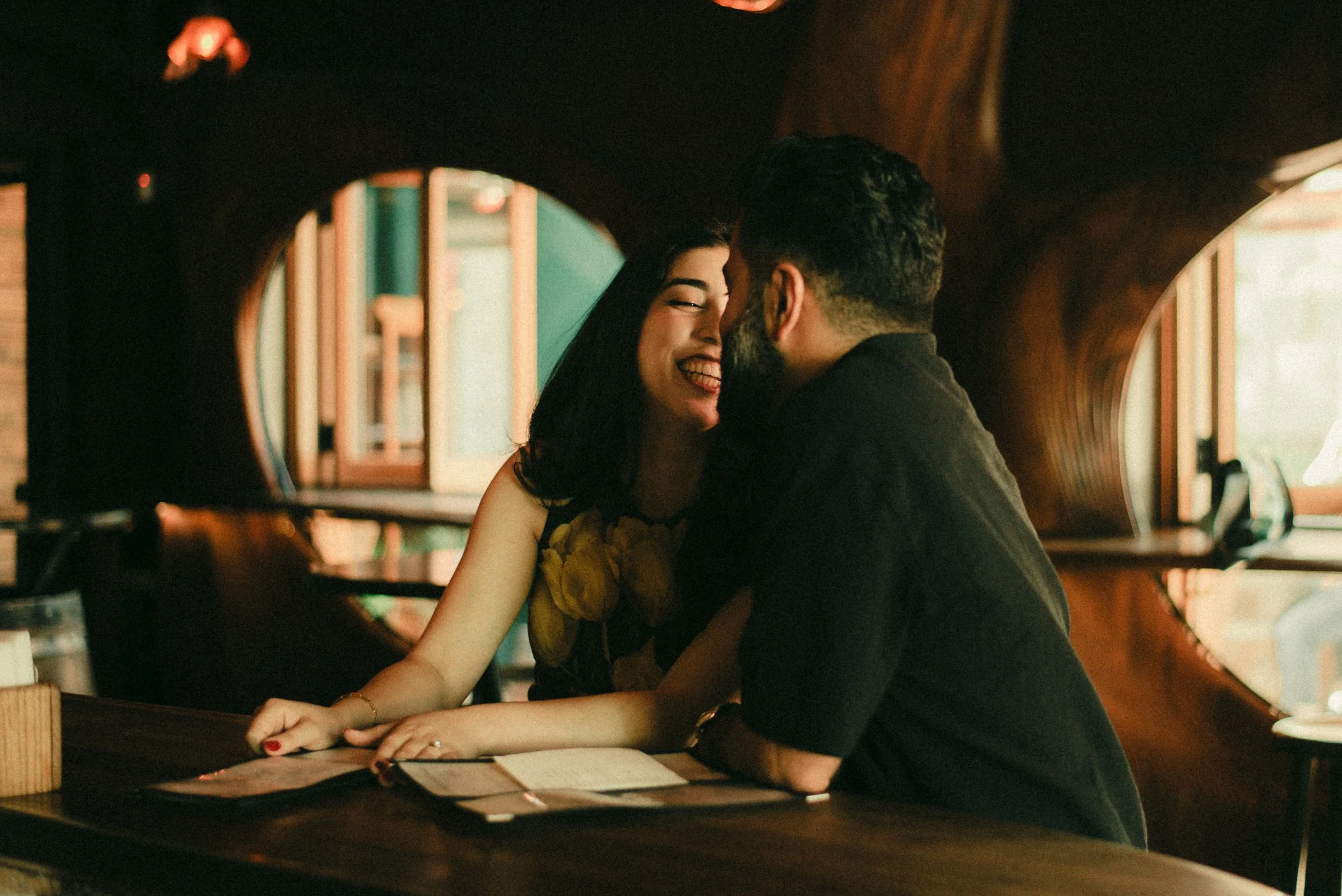 A man and woman sharing a close, joyful moment at a restaurant table, smiling and leaning toward each other.